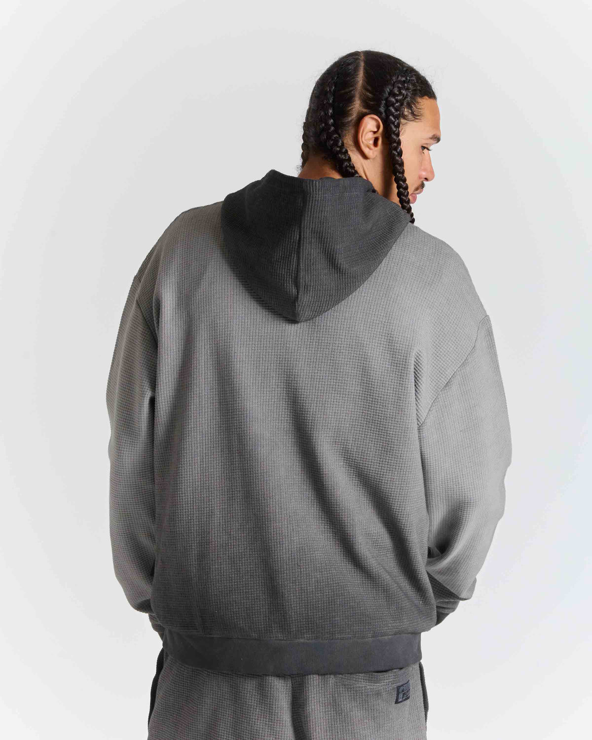 A man wearing a gray textured hooded sweatshirt and matching sweatpants, shown from the back against a light gray studio background.