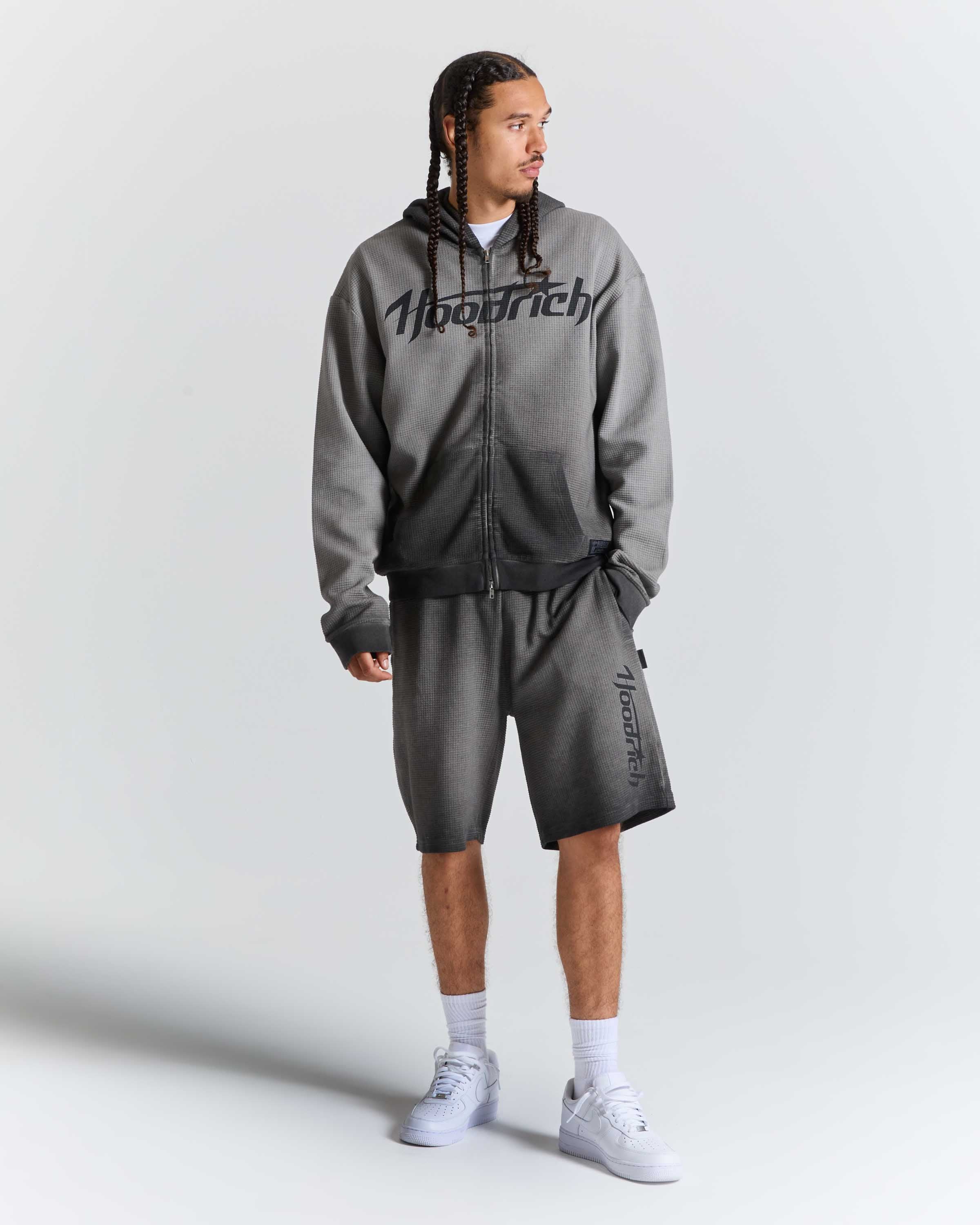 A man modeling a gray zip-up hoodie and matching gray shorts with black 'Hoodrich' branding, styled with white socks and white sneakers against a light gray studio background.