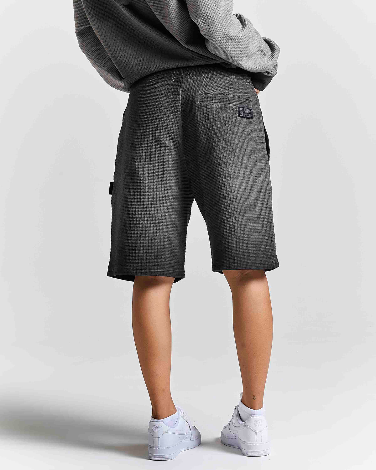 A person shown from the back wearing dark gray textured shorts, a gray sweatshirt, and white sneakers against a light gray studio background.