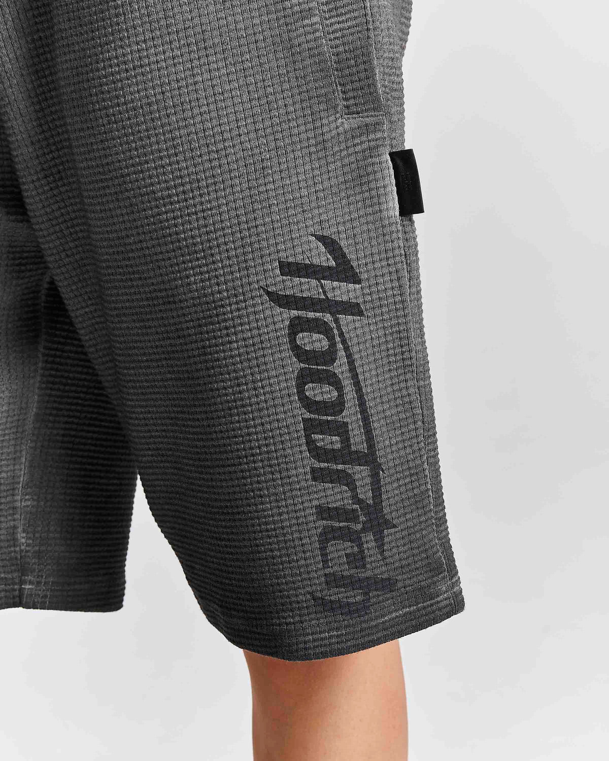 Close-up of dark gray waffle-knit shorts with a large black vertical 'hooded' logo printed on the side, worn by a model against a light gray background.