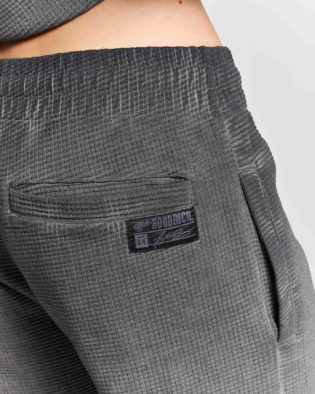 Close-up of dark gray ribbed knit pants showing a side pocket with a black and white HOODRICH brand patch, worn by a model.