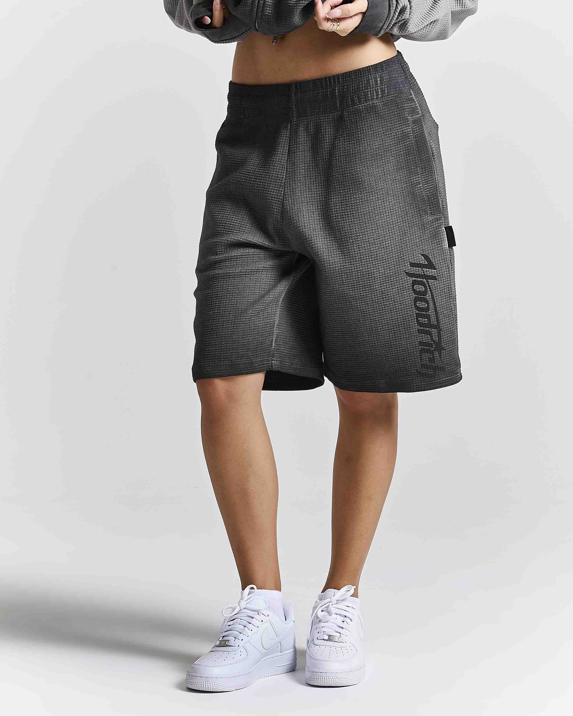 A person wearing dark gray textured athletic shorts with a vertical black 'Mood' logo on the side, paired with white sneakers and a gray cropped jacket, shown from the waist down against a light gray studio background.