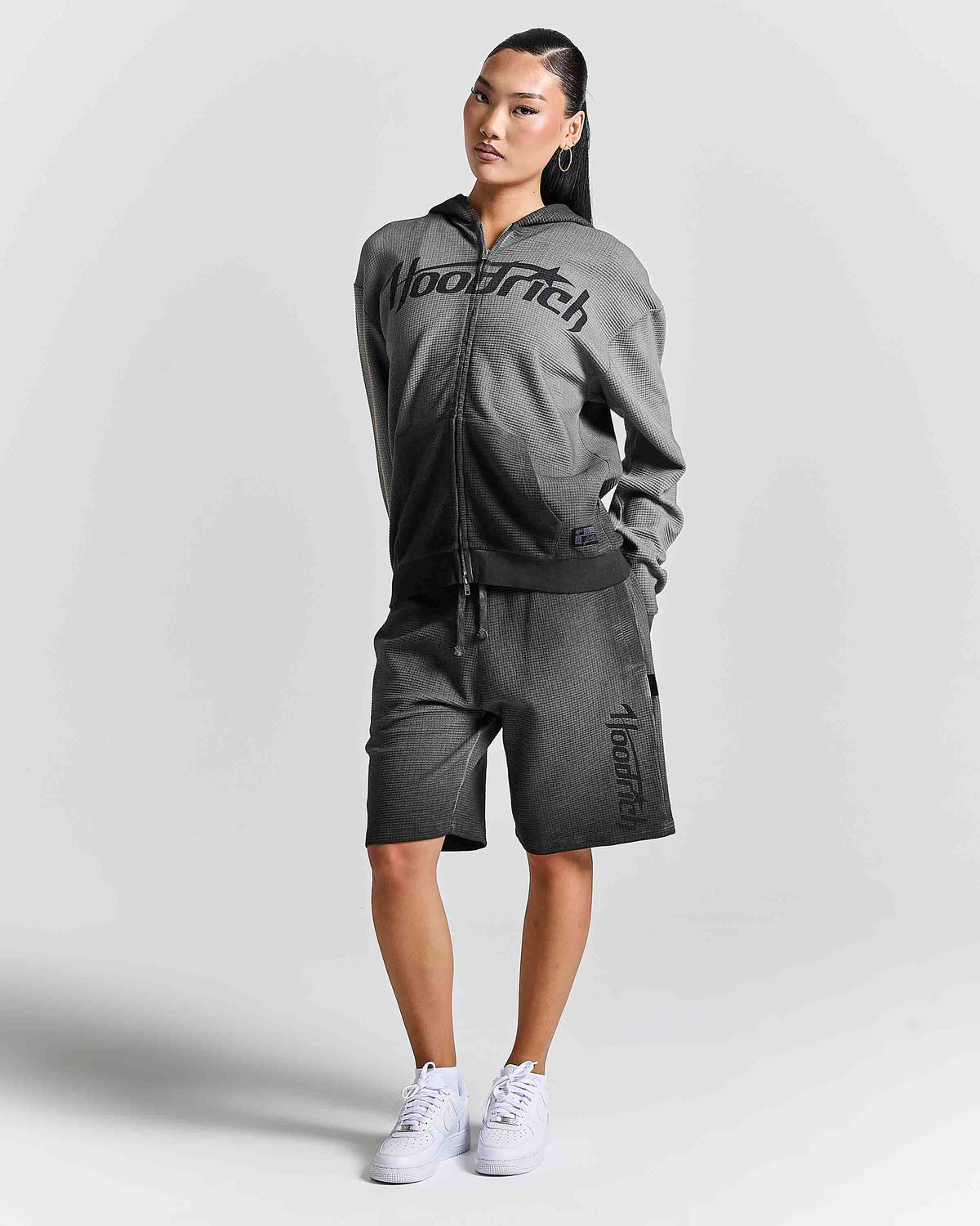 A woman modeling a dark gray two-piece tracksuit featuring a zip-up hoodie with 'HOODRICH' printed across the chest and matching shorts with 'HOODRich' printed down the leg, paired with white sneakers against a light gray studio background.