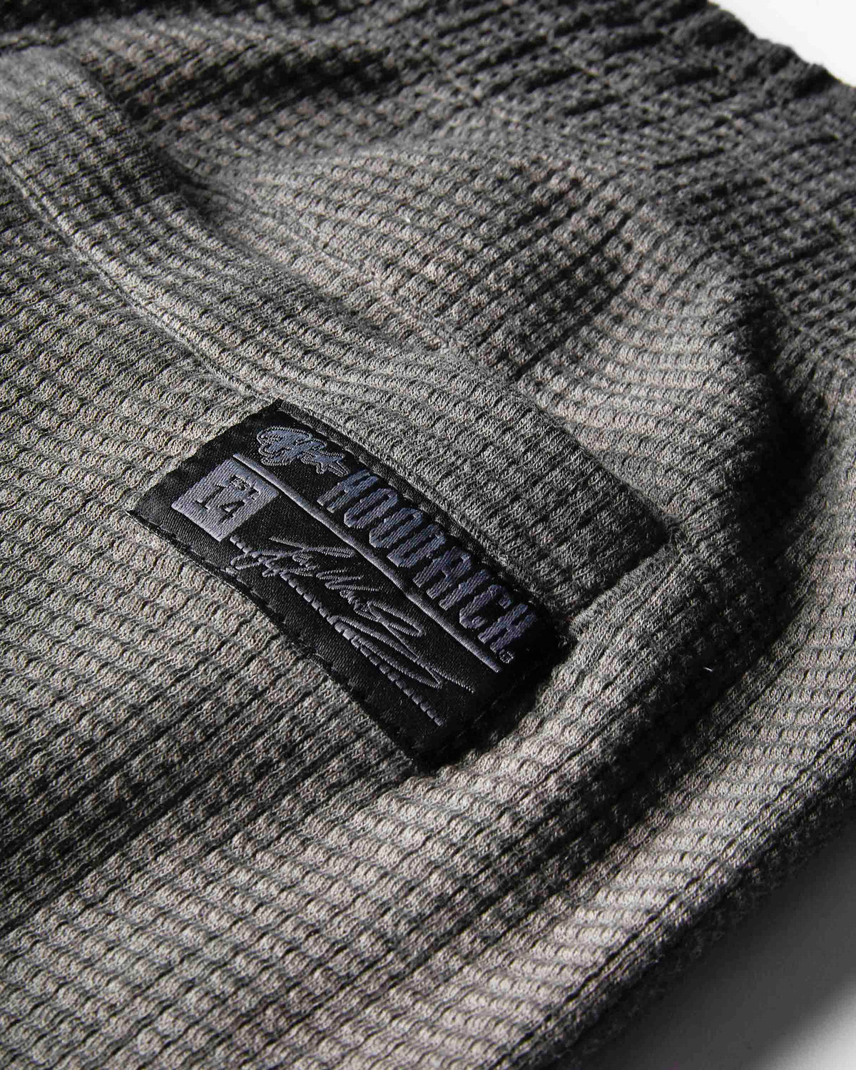 Close-up of a dark gray waffle-knit fabric with a black embroidered patch reading 'hoodrich' and on a white background.