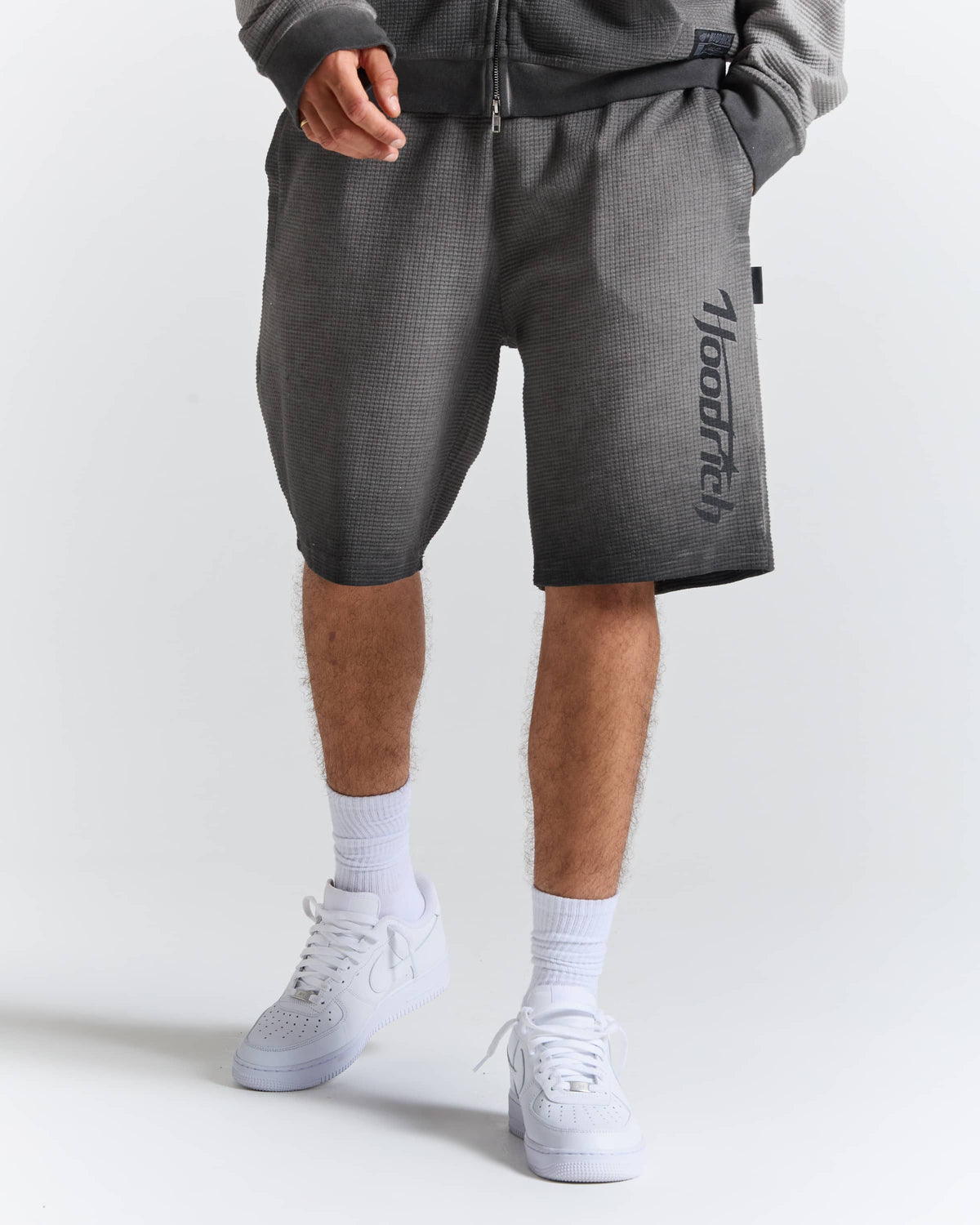 A person wearing dark gray textured athletic shorts with a vertical black 'hoodlits' logo on the left leg, styled with white crew socks and white sneakers against a light gray studio background.