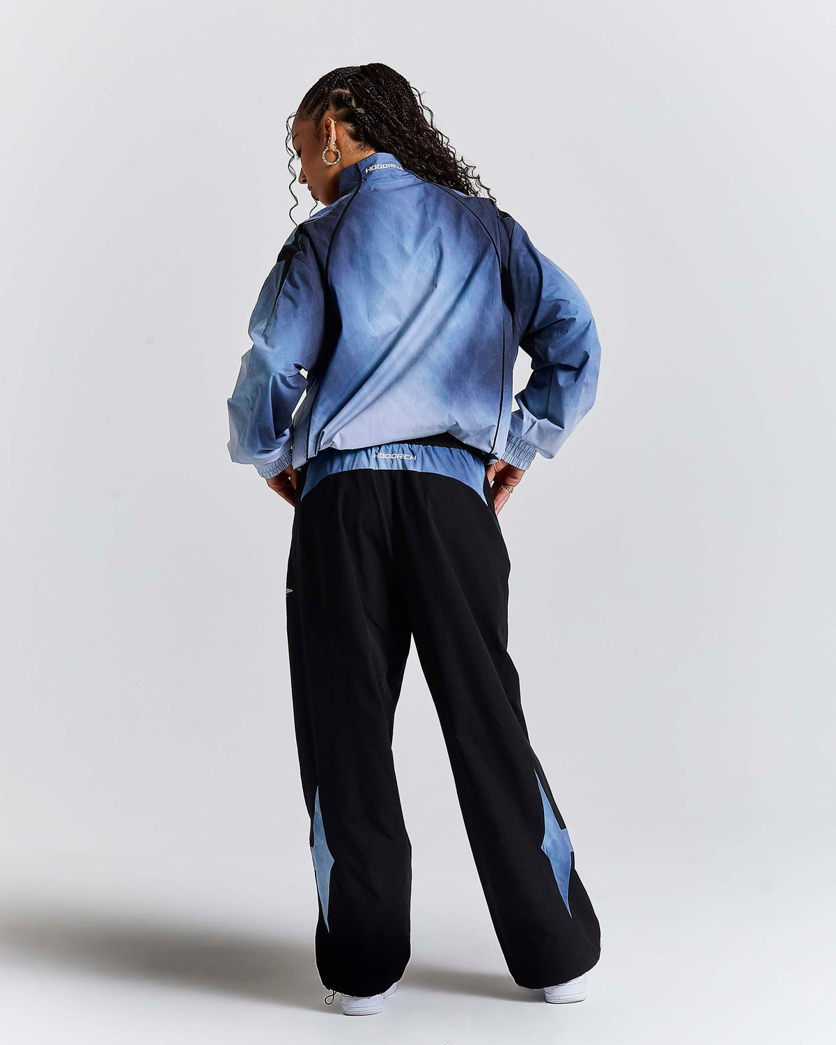 A model is shown from behind wearing a blue tie-dye bomber jacket and black wide-leg sweatpants with blue star accents on the lower legs, set against a plain light gray studio background.