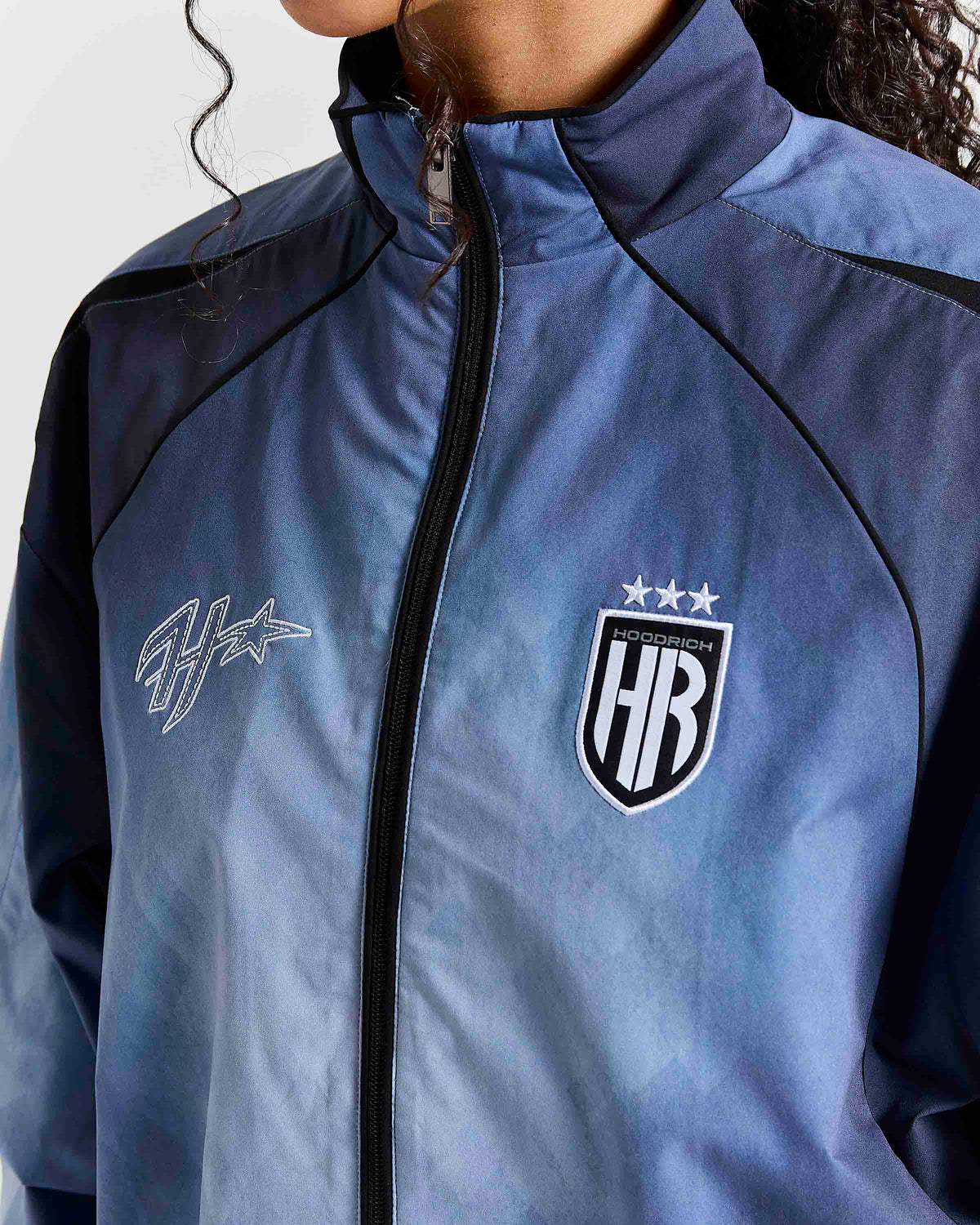 A close-up of a person wearing a blue and navy athletic jacket with black zippers and logos, including a shield crest with three stars and a stylized script logo on the chest.