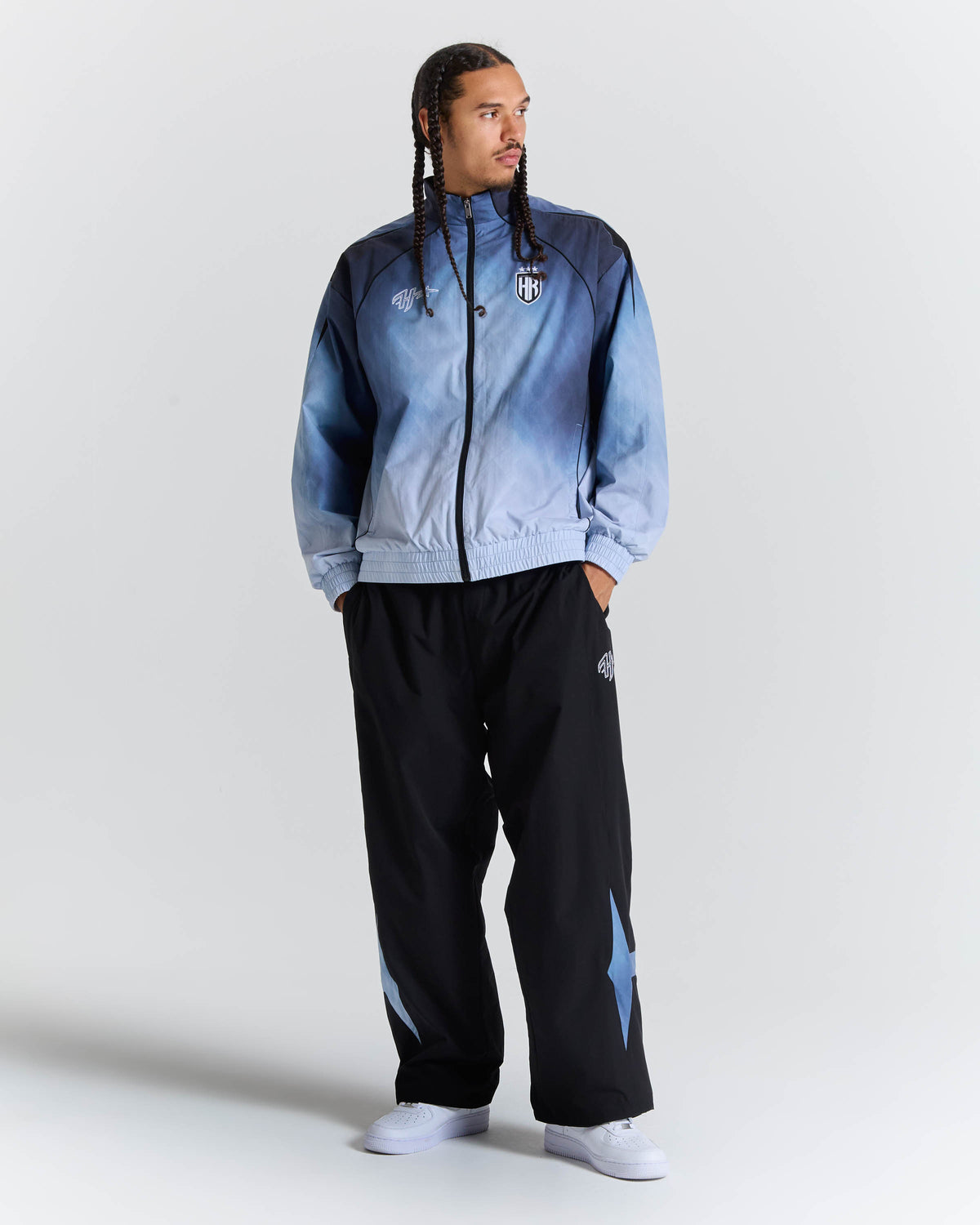 A man modeling a light blue and navy gradient zip-up jacket with black track pants featuring light blue star details, set against a plain light gray studio background.