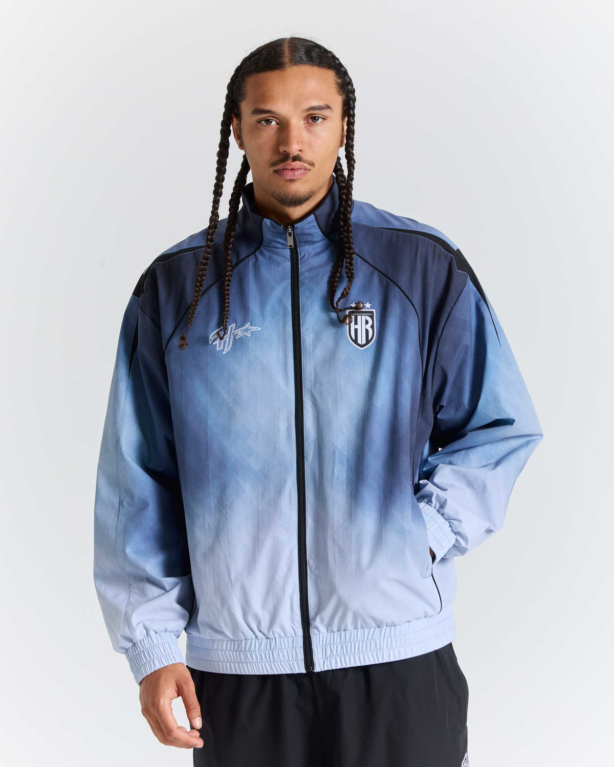 A man wearing a light blue and navy gradient zip-up jacket with a shield crest on the left chest, photographed against a plain light gray studio background.