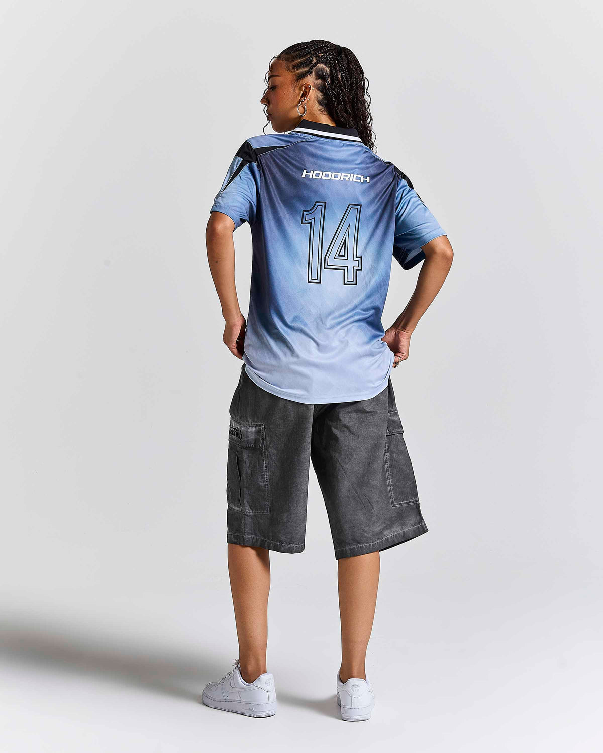 A child model shown from the back wearing a light blue gradient soccer jersey with 'HOODRICH' and the number '14' on the back, paired with dark gray cargo shorts and white sneakers, against a light gray studio background.