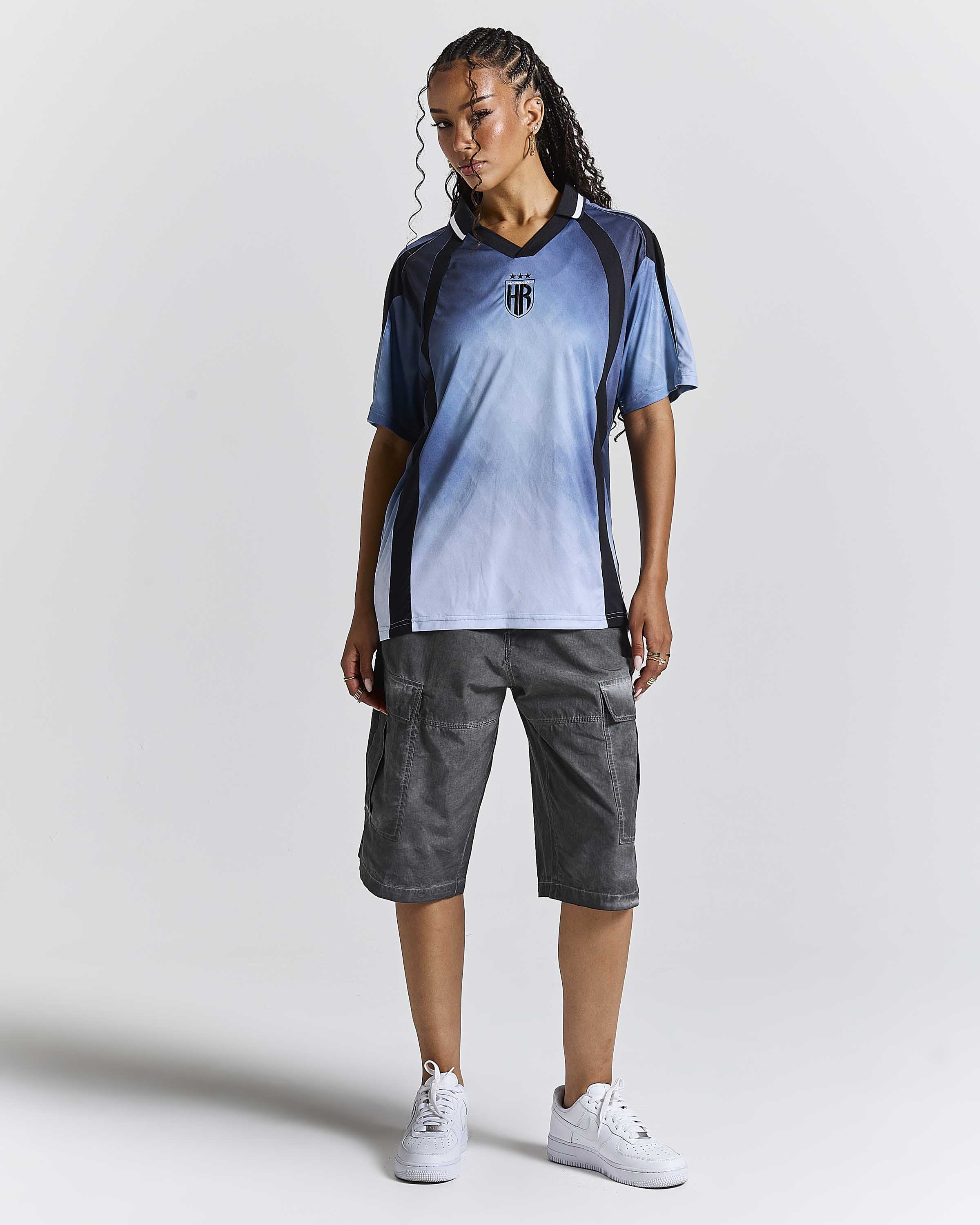 A woman modeling a blue and white gradient soccer jersey with black shoulder panels, paired with dark gray cargo shorts and white sneakers, against a light gray studio background.
