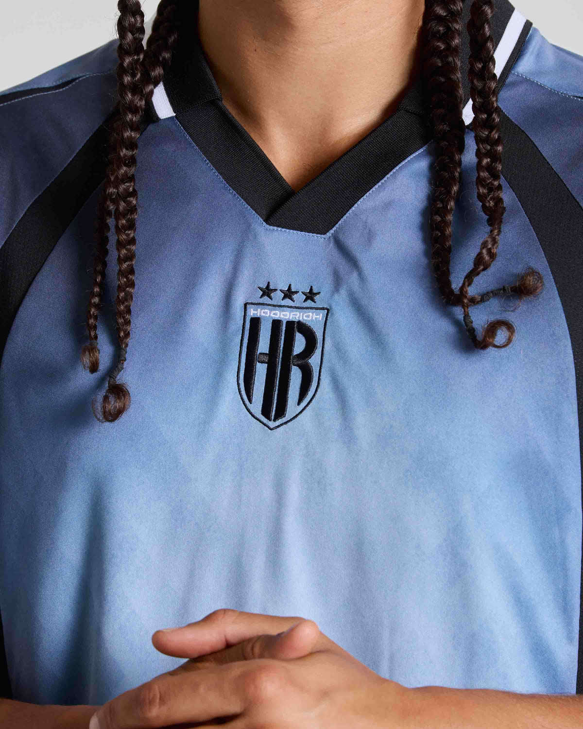 A close-up of a person wearing a light blue soccer jersey with black trim, featuring the 'HOODRICH' crest on the chest.