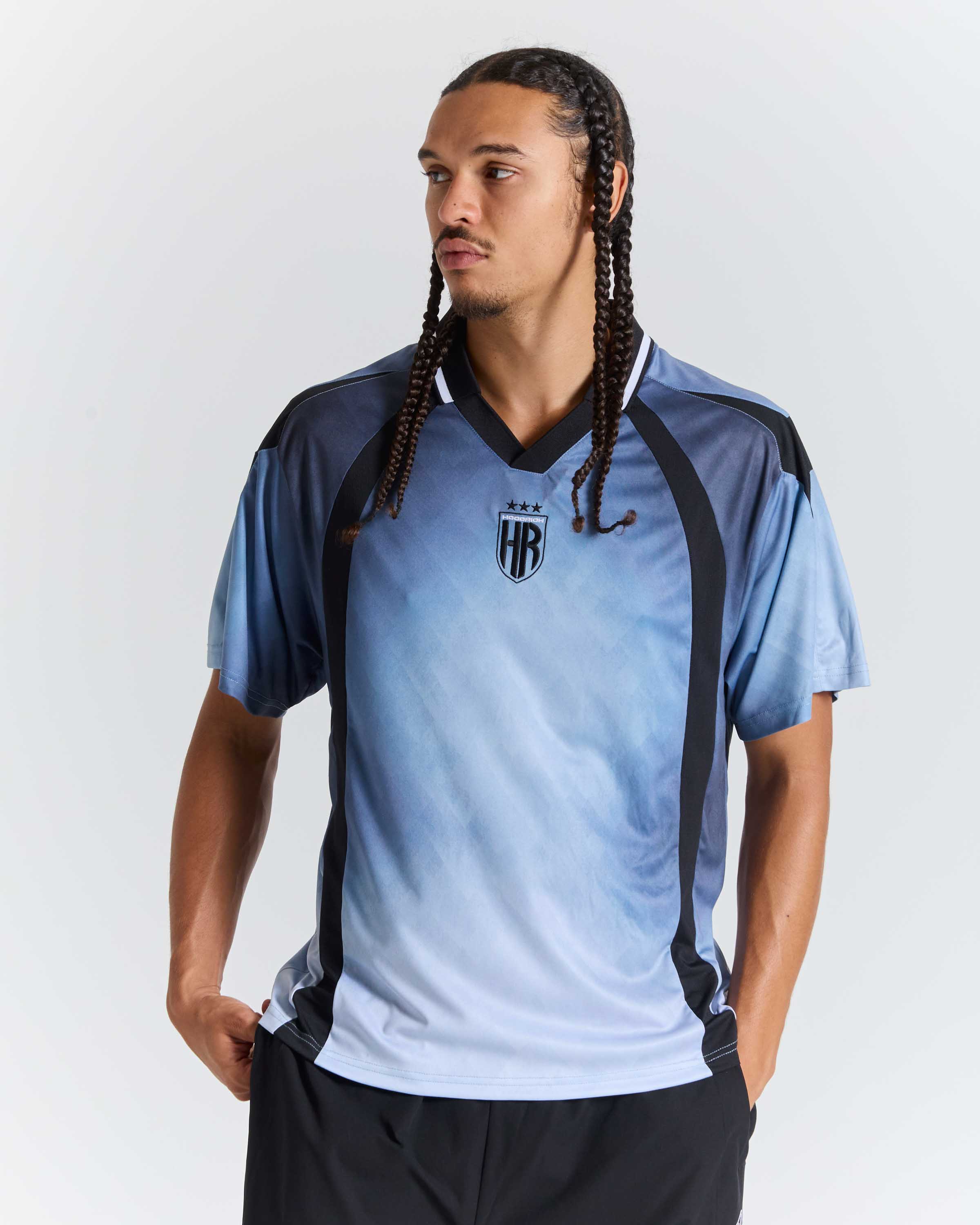 A man modeling a light blue and navy soccer jersey with black side panels and a central crest, set against a plain light gray background.