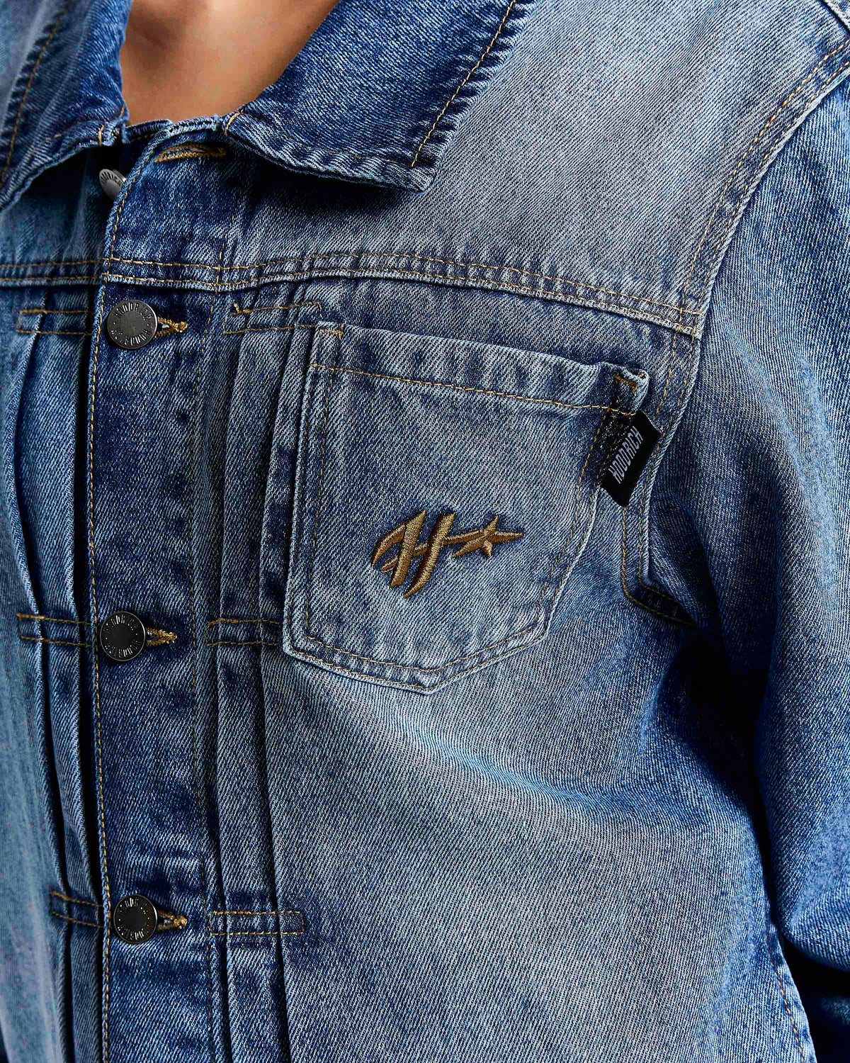 Close-up of a blue denim jacket featuring a chest pocket with a gold embroidered logo and dark metal snap buttons.