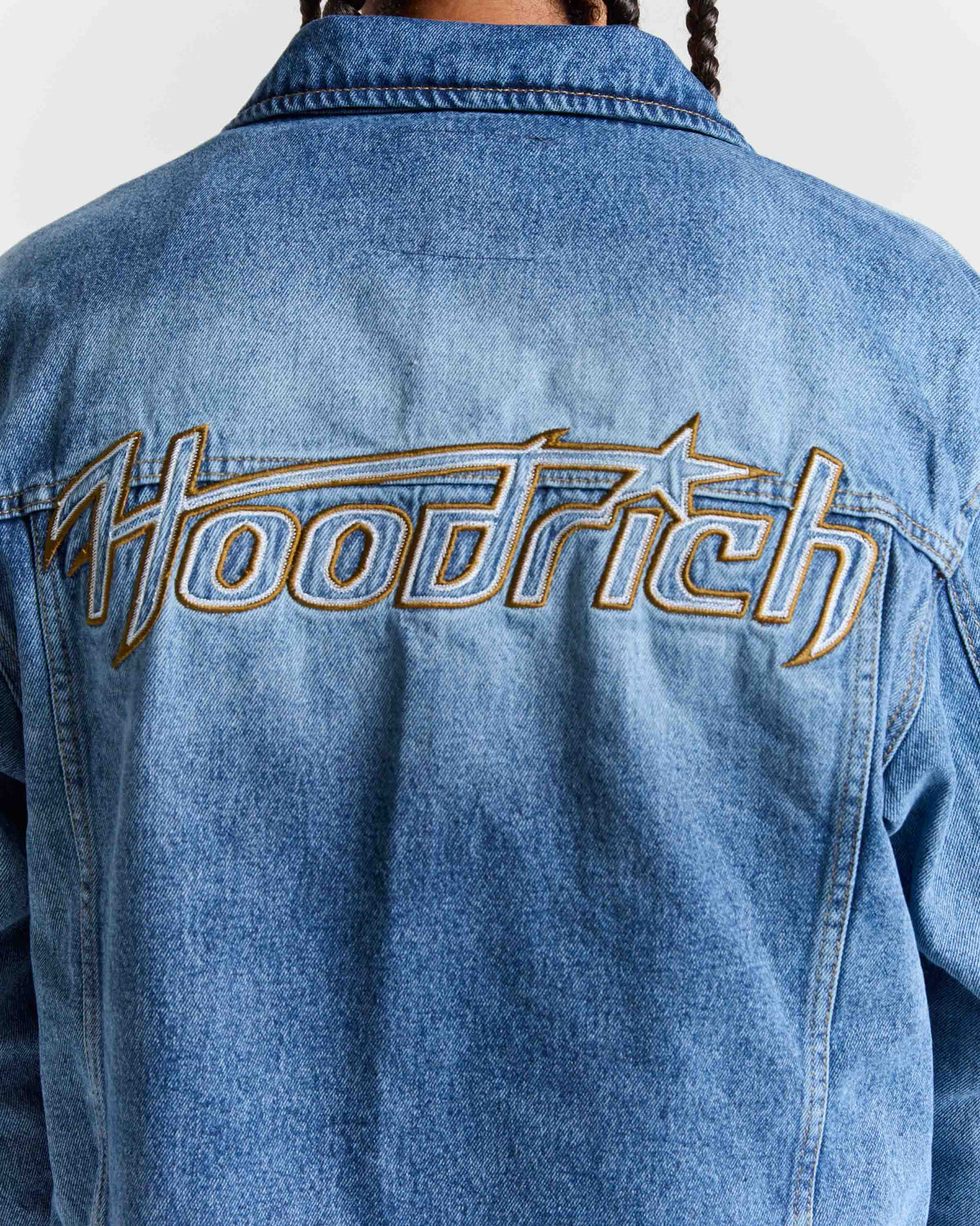 Close-up back view of a person wearing a blue denim jacket with a large embroidered 'Hoodrich' logo across the upper back in gold and light blue.