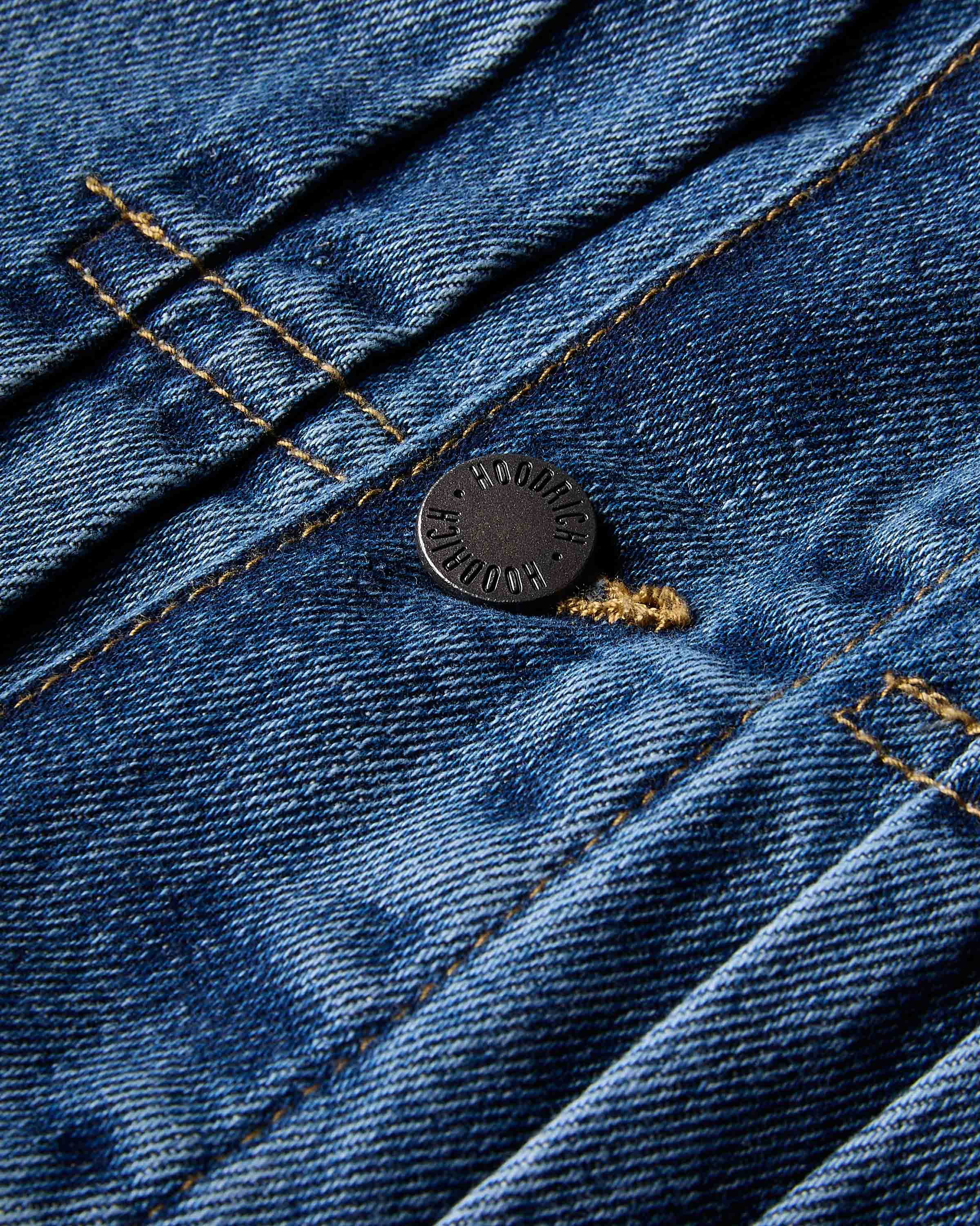 Close-up of a dark metal button on a blue denim garment, showing the textured fabric weave and contrast stitching.