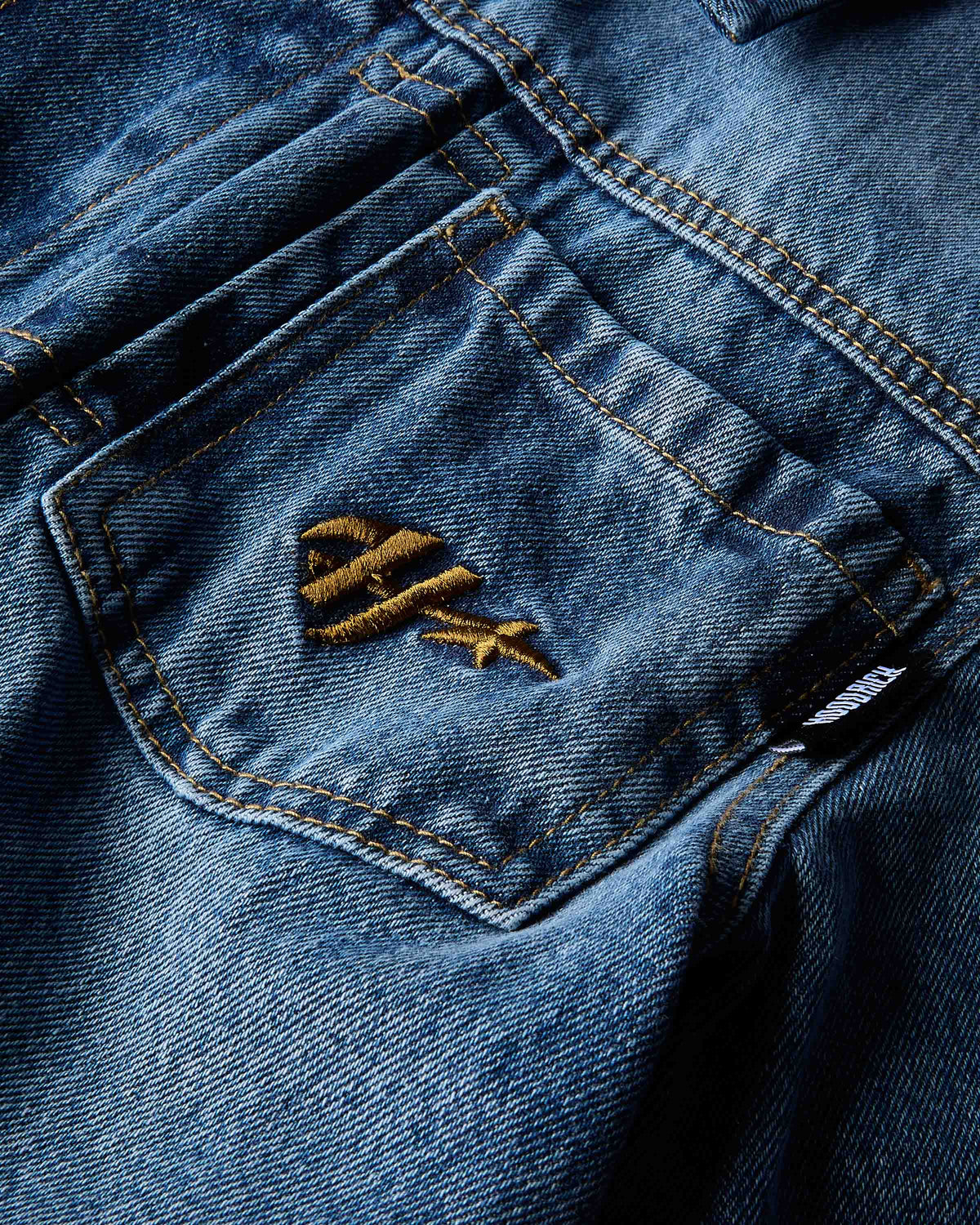 Close-up of a blue denim jacket pocket featuring a gold embroidered logo and contrast gold stitching.