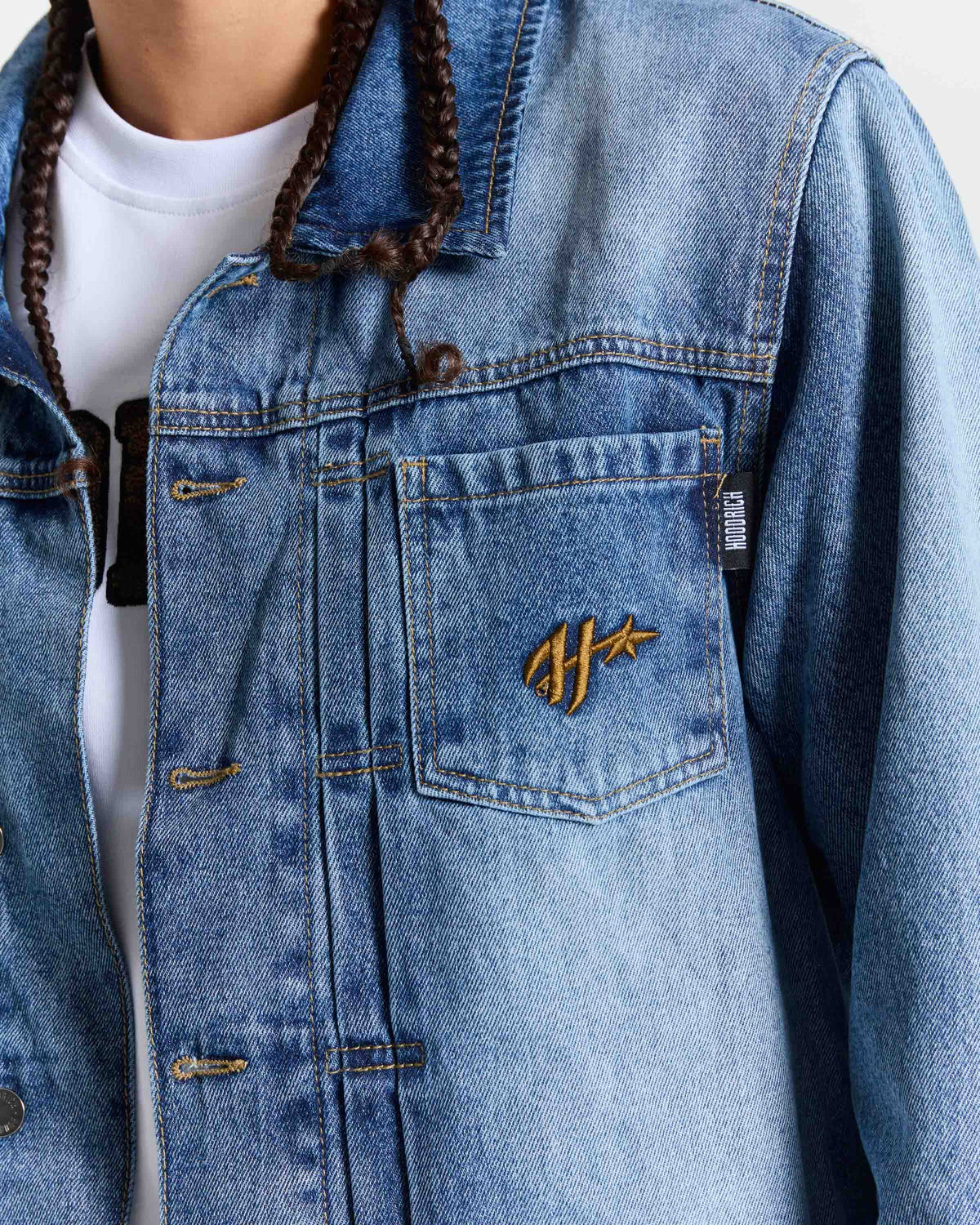 A close-up of a person wearing a light blue denim jacket over a white t-shirt. The jacket features a chest pocket with a gold embroidered logo and a small black brand tag.