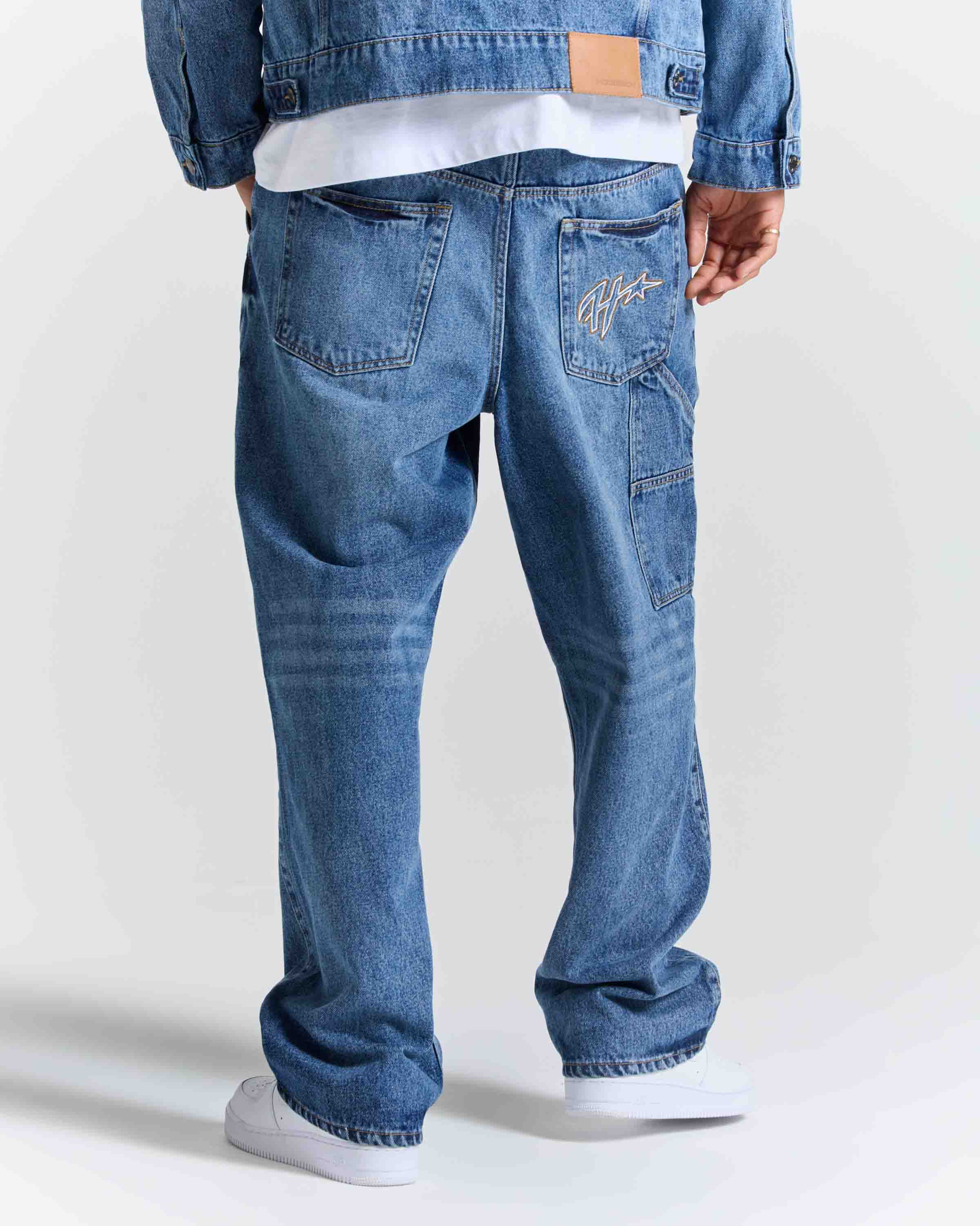 Rear view of a person wearing blue denim jeans with a white t-shirt and white sneakers, showing the back pockets and signature embroidery on the right pocket.