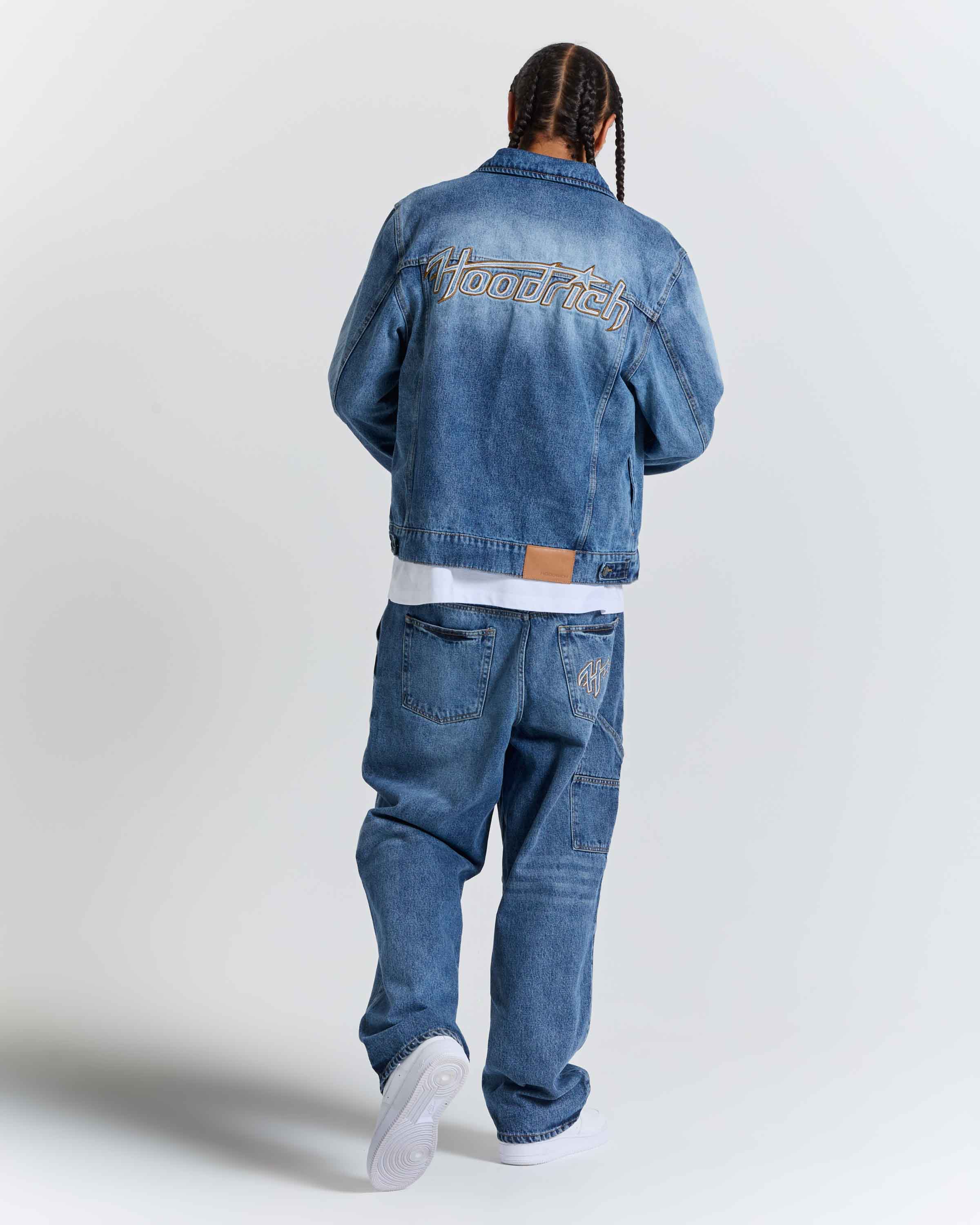 A man is shown from behind wearing a blue denim jacket with 'HoodRich' embroidered on the back, matching blue jeans, a white t-shirt, and white sneakers, set against a plain light gray studio background.