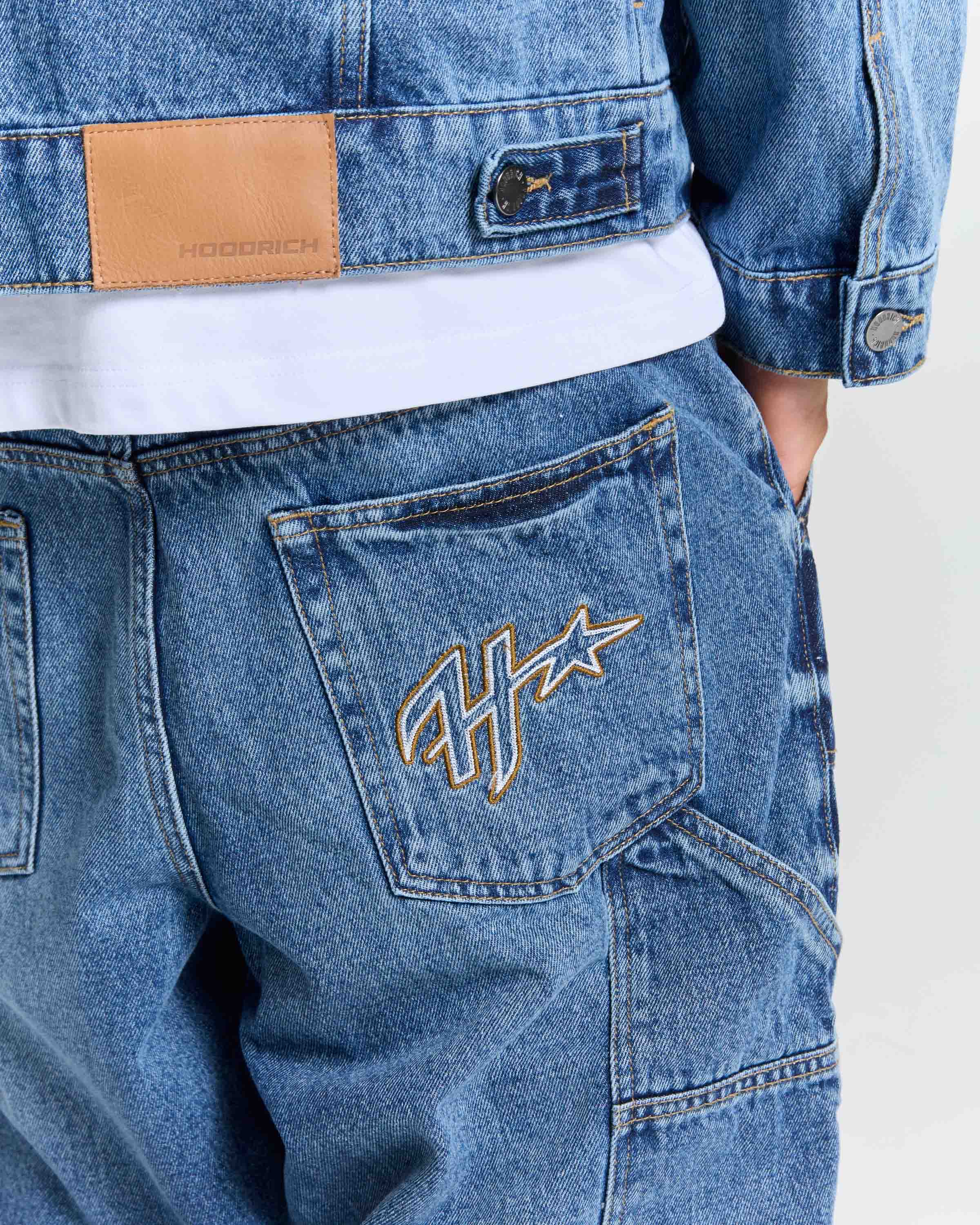 Close-up of a person wearing blue denim jeans with a white t-shirt underneath and a matching blue denim jacket. The back pocket of the jeans features a gold embroidered monogram logo, and a tan leather brand patch is visible on the jacket.