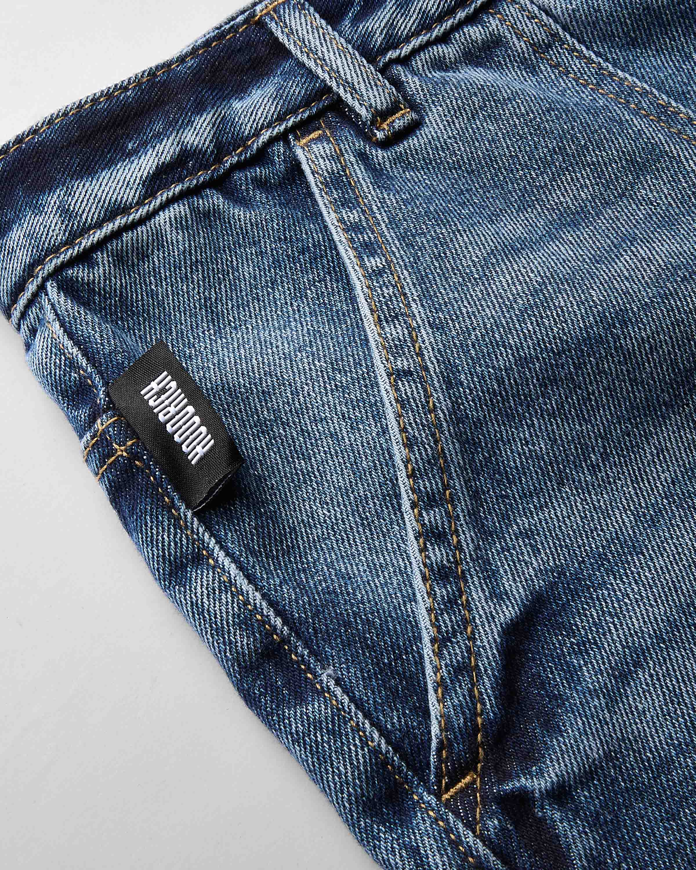 Close-up of blue denim jeans showing the back pocket, stitching, and a black fabric brand tab that reads 'HOODRICH'.