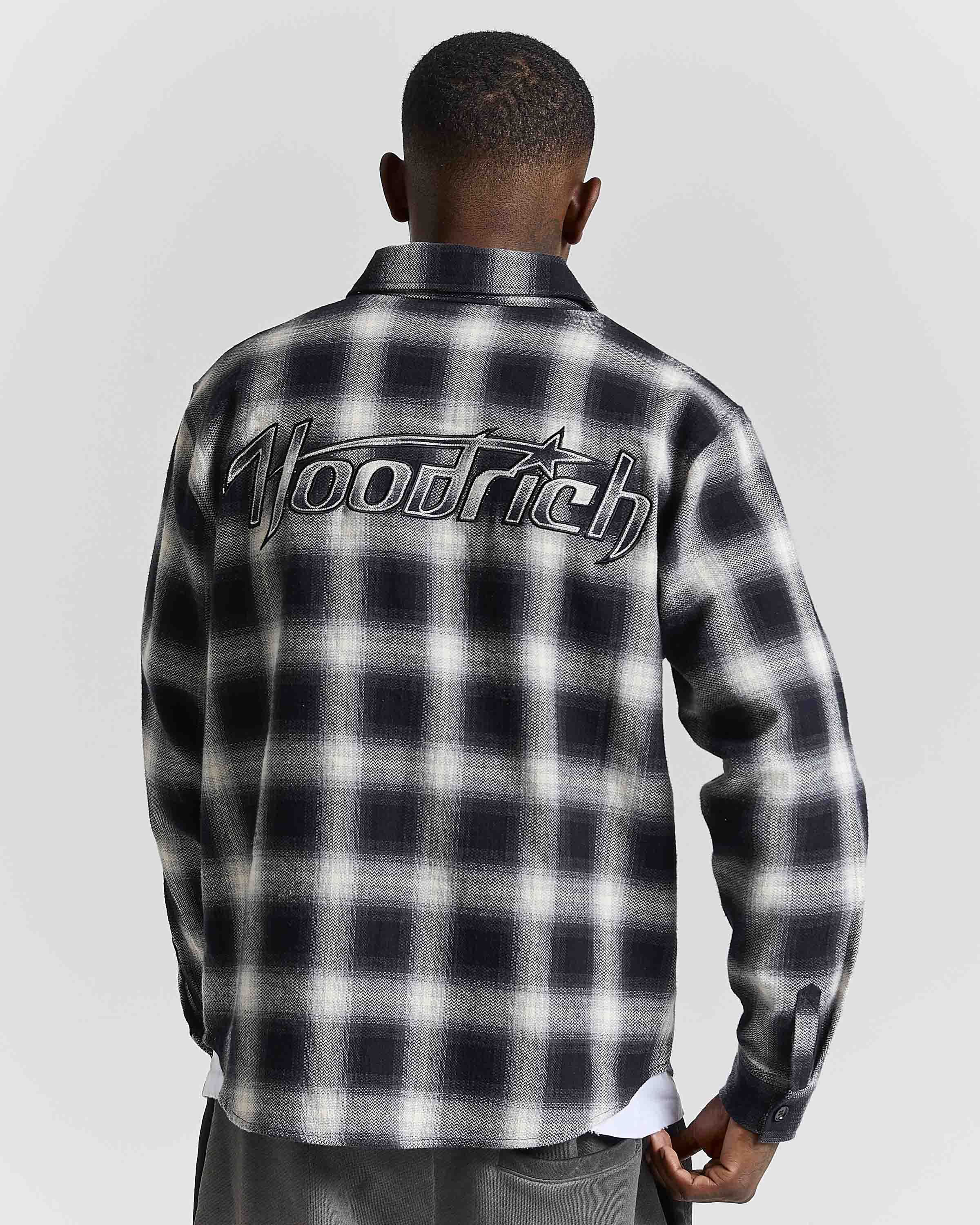 A man viewed from behind wearing a black and white plaid button-up shirt with a large 'Hoodrich' logo across the upper back, paired with gray pants against a light gray studio background.