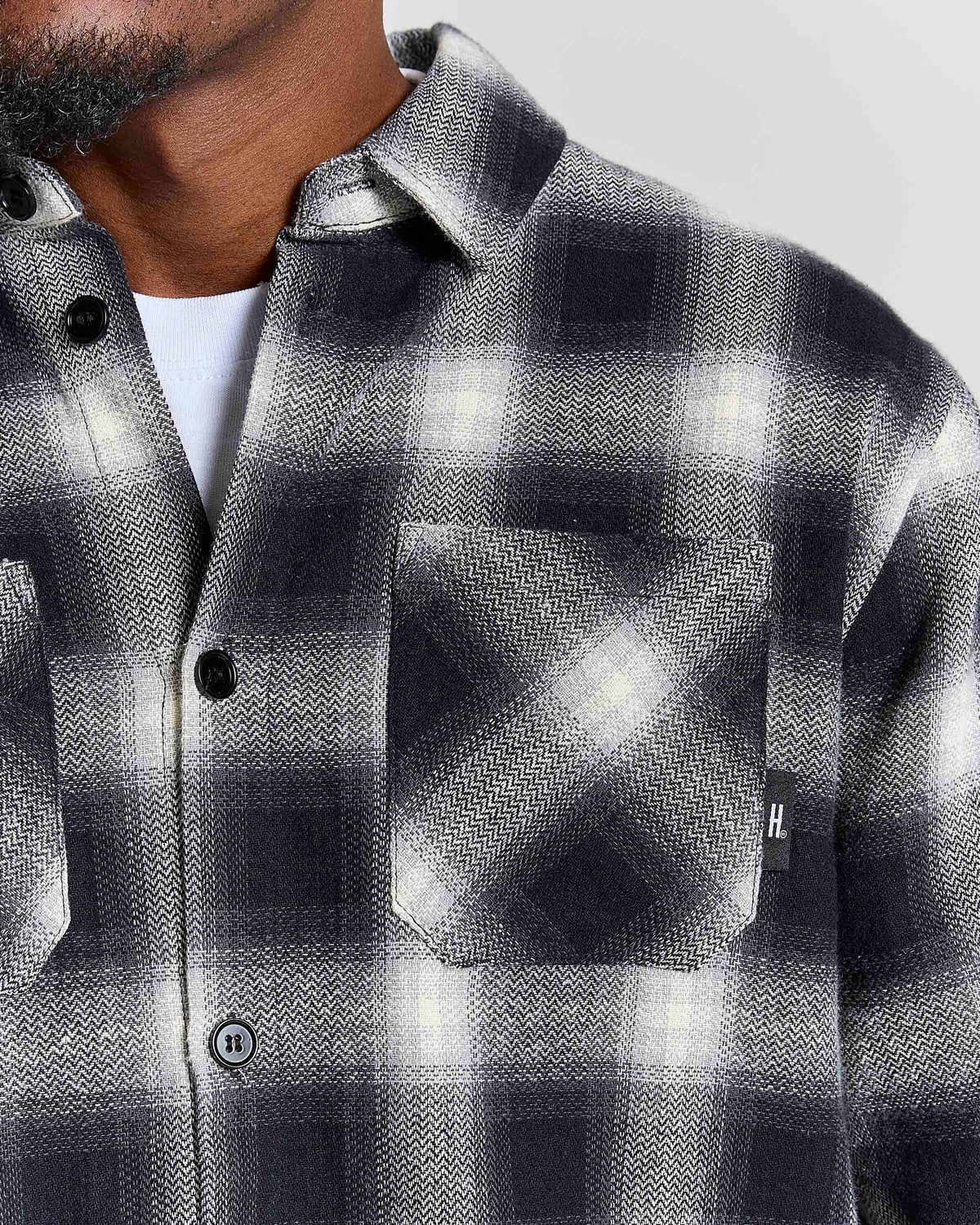 Close-up of a man wearing a black, white, and gray plaid button-up shirt with a chest pocket, layered over a white undershirt.