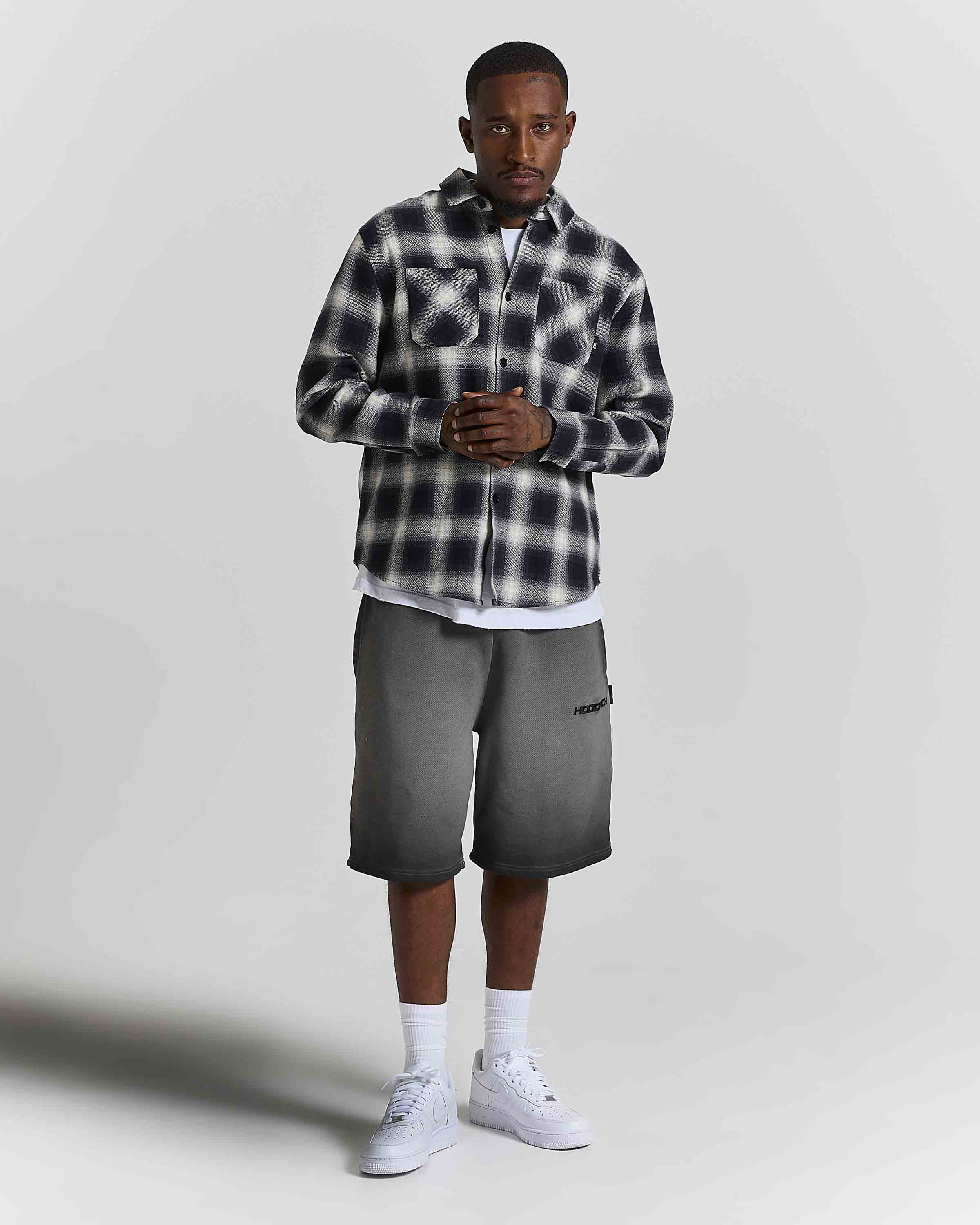 A man modeling a black and white plaid button-up shirt, dark gray shorts, white socks, and white sneakers against a light gray studio background.