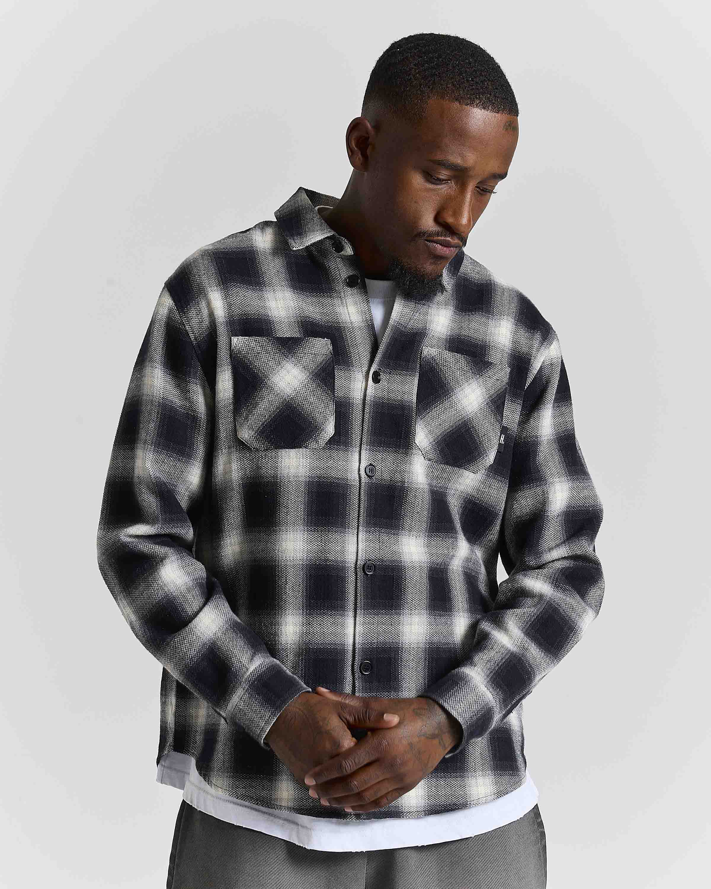 A man wearing a black and white plaid button-up flannel shirt over a white undershirt, standing against a light gray studio background.