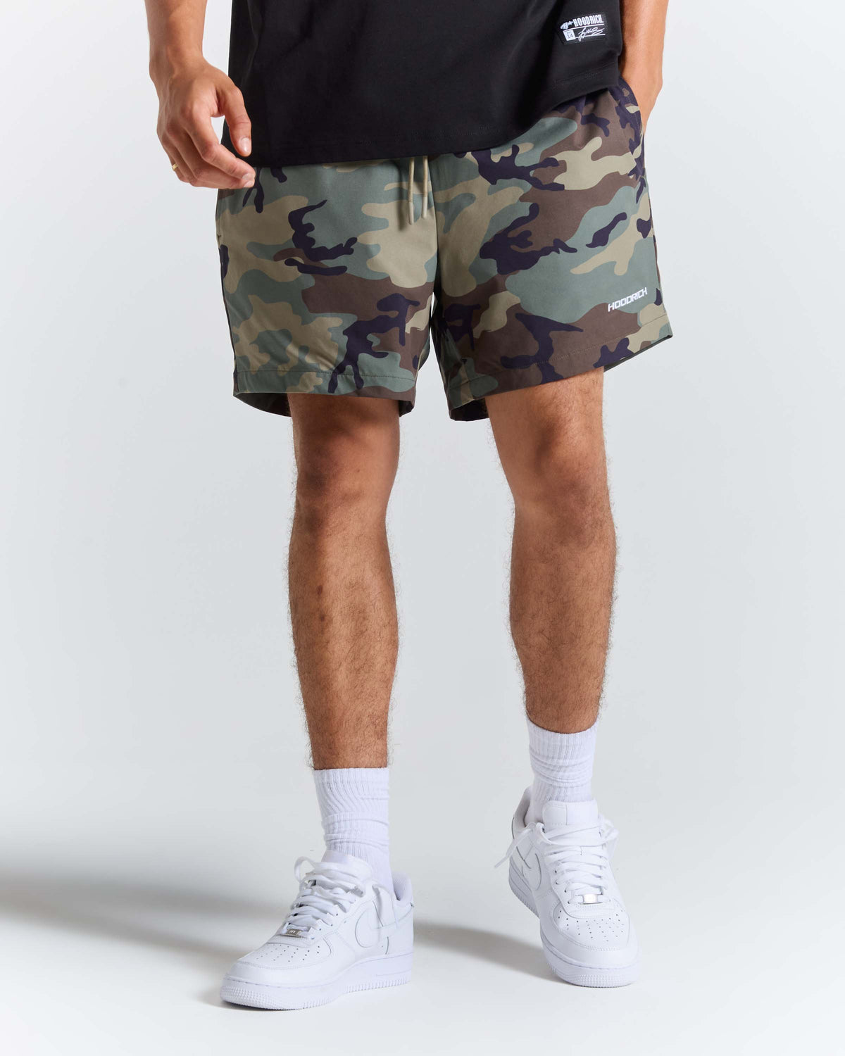 A person wearing camouflage athletic shorts, a black t-shirt, white socks, and white sneakers, shown from the waist down on a light gray studio background.