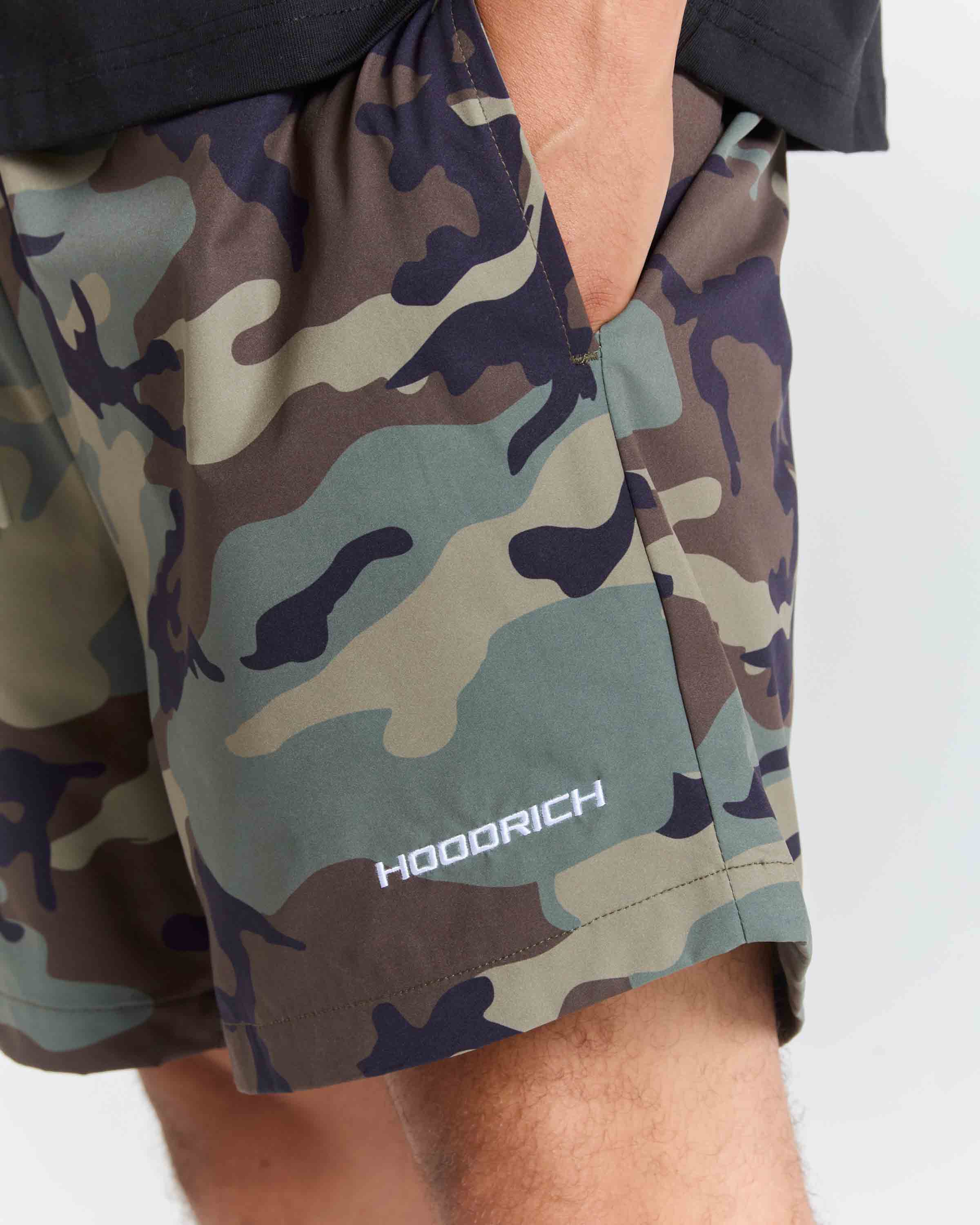 Close-up of a person wearing camouflage shorts with a visible HOODRICH logo near the hem, against a light gray background.