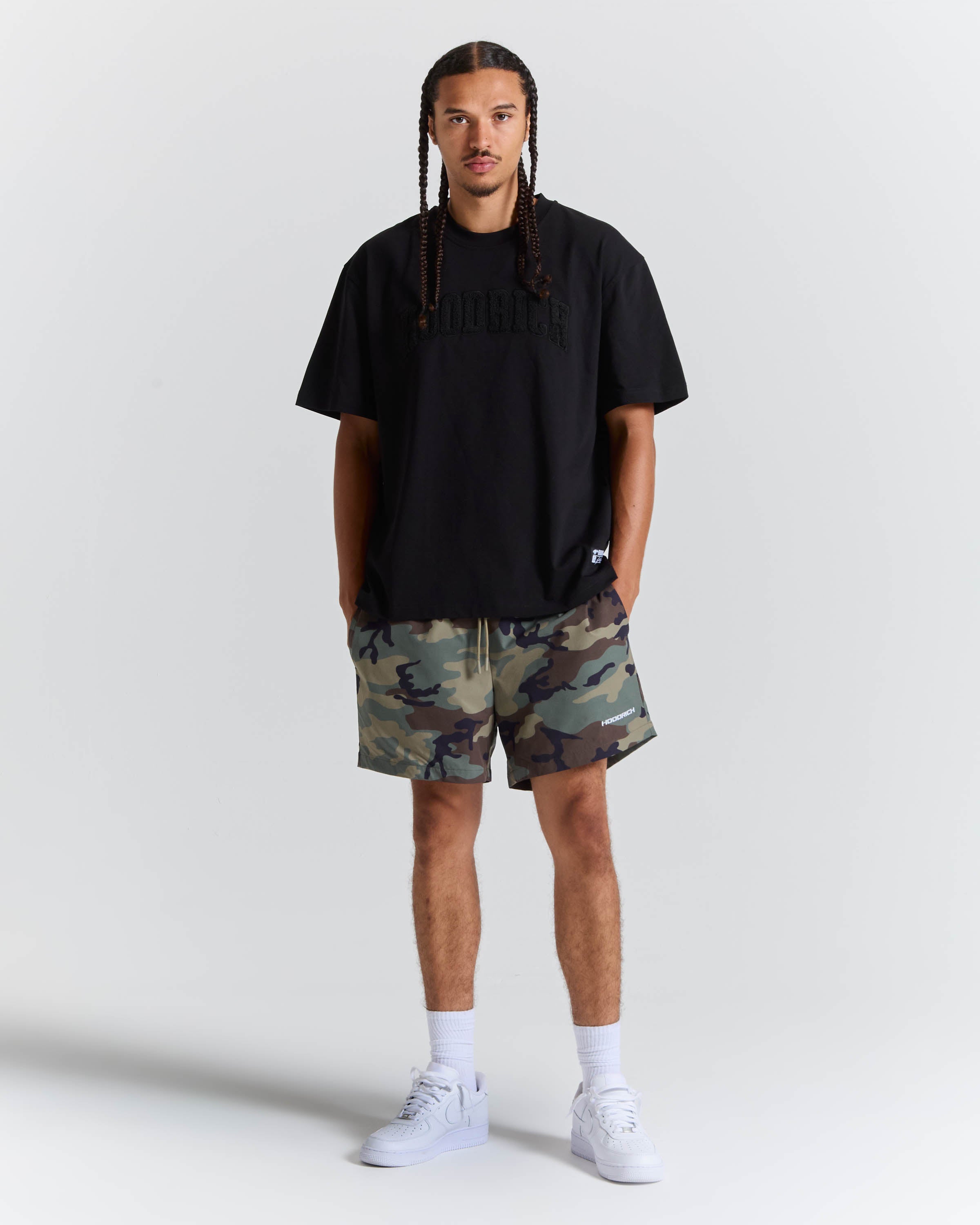 A man modeling a black t-shirt and camouflage shorts against a light gray studio background.