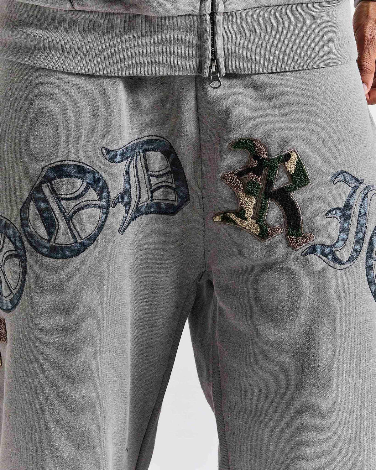 Close-up of light gray sweatpants with large decorative embroidered lettering across the thighs and a camouflage patch on the right thigh.
