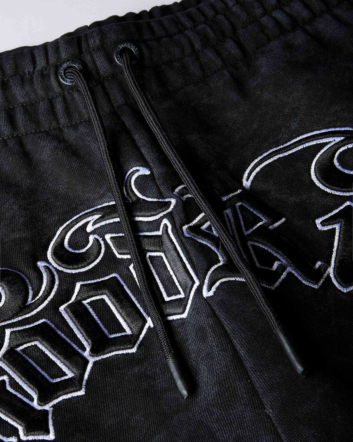 Close-up of black drawstring shorts with large white and blue outlined gothic-style lettering across the front.