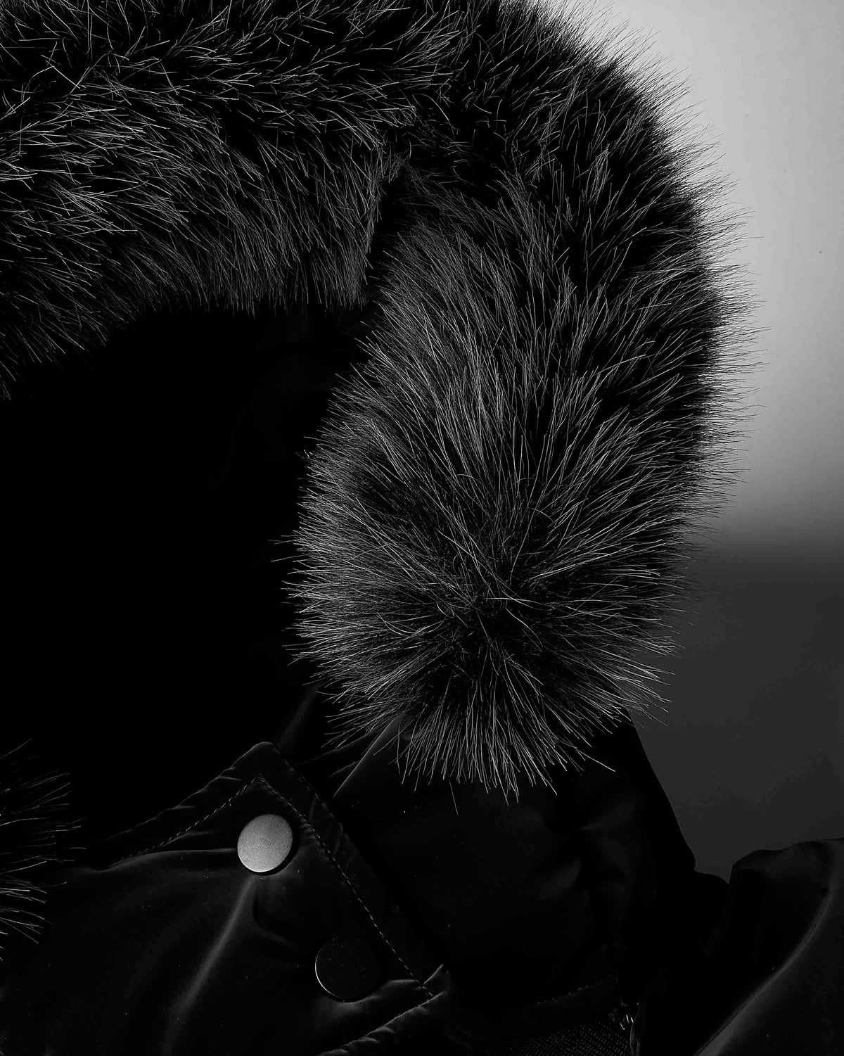 Close-up of a black winter jacket with a large dark fur hood trim and a silver snap button, shown against a dark gray background.