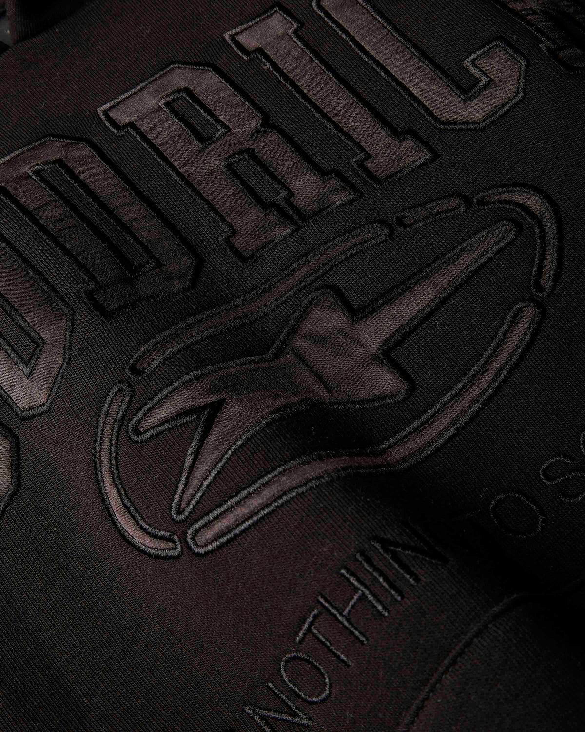 Close-up of a dark embroidered graphic on fabric, showing large raised letters and a stylized logo in a very dark brown or charcoal tone against a black background.