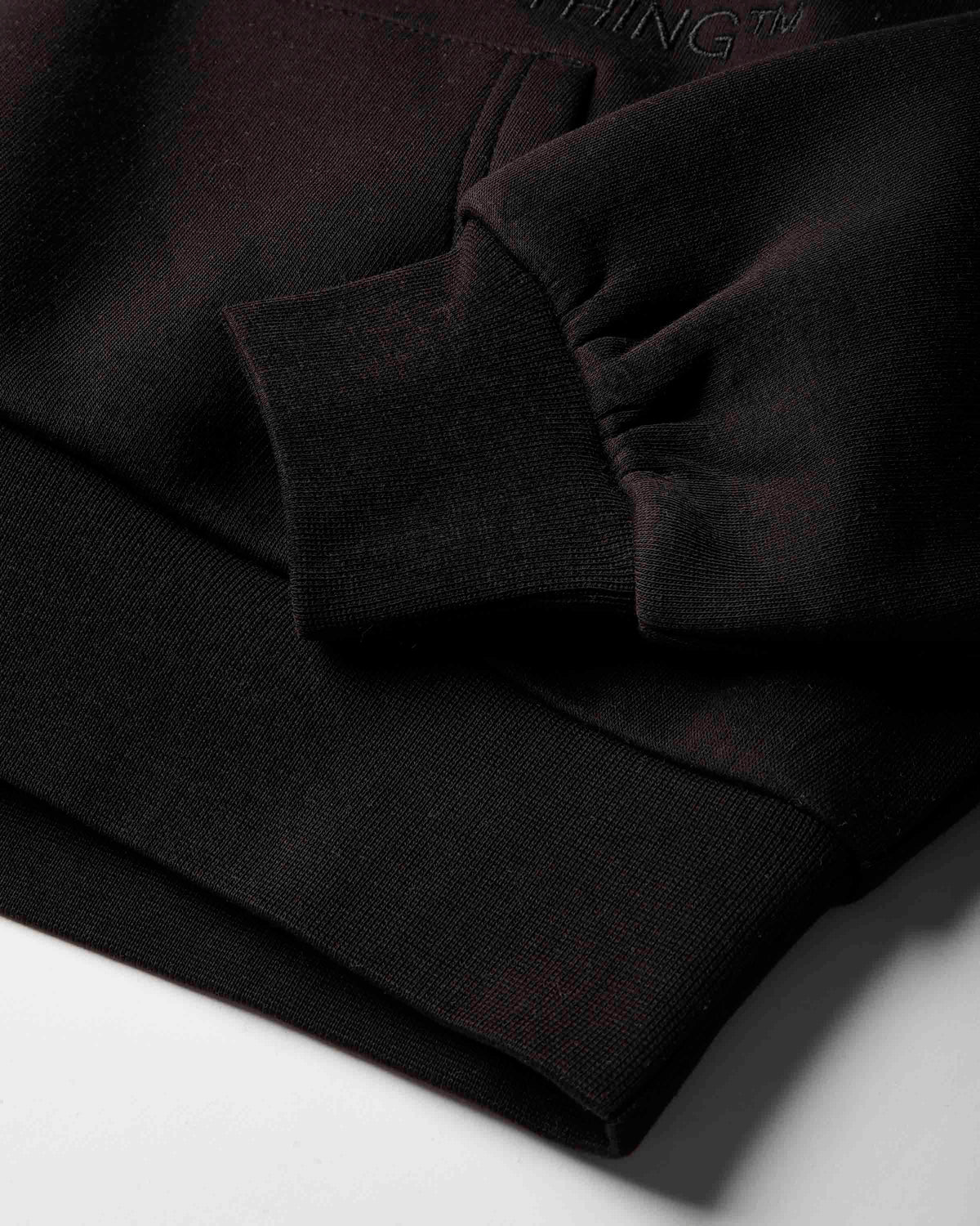 Close-up of a black ribbed knit garment, likely a sweatshirt or hoodie, showing the cuff, hem, and a small tonal embroidered brand mark reading NOTHING on a white surface.