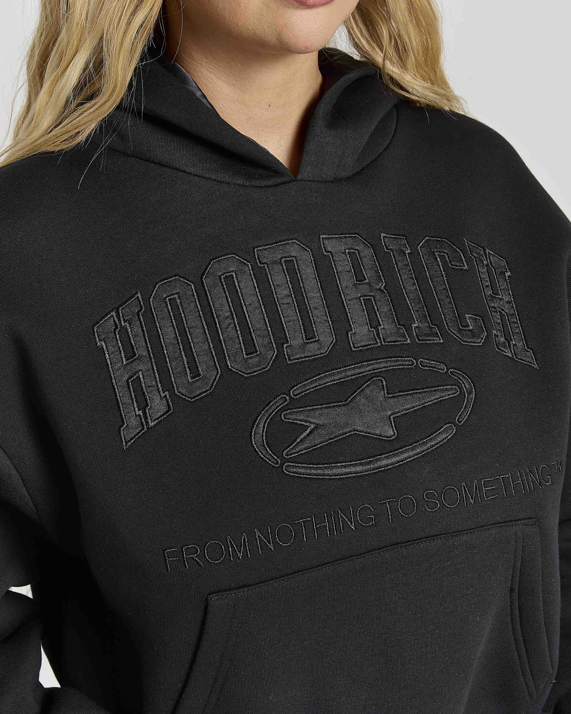 Close-up of a person wearing a black hoodie with tonal dark gray 'HOODRICH' lettering and a star logo on the chest, plus smaller text reading 'FROM NOTHING TO SOMETHING'.