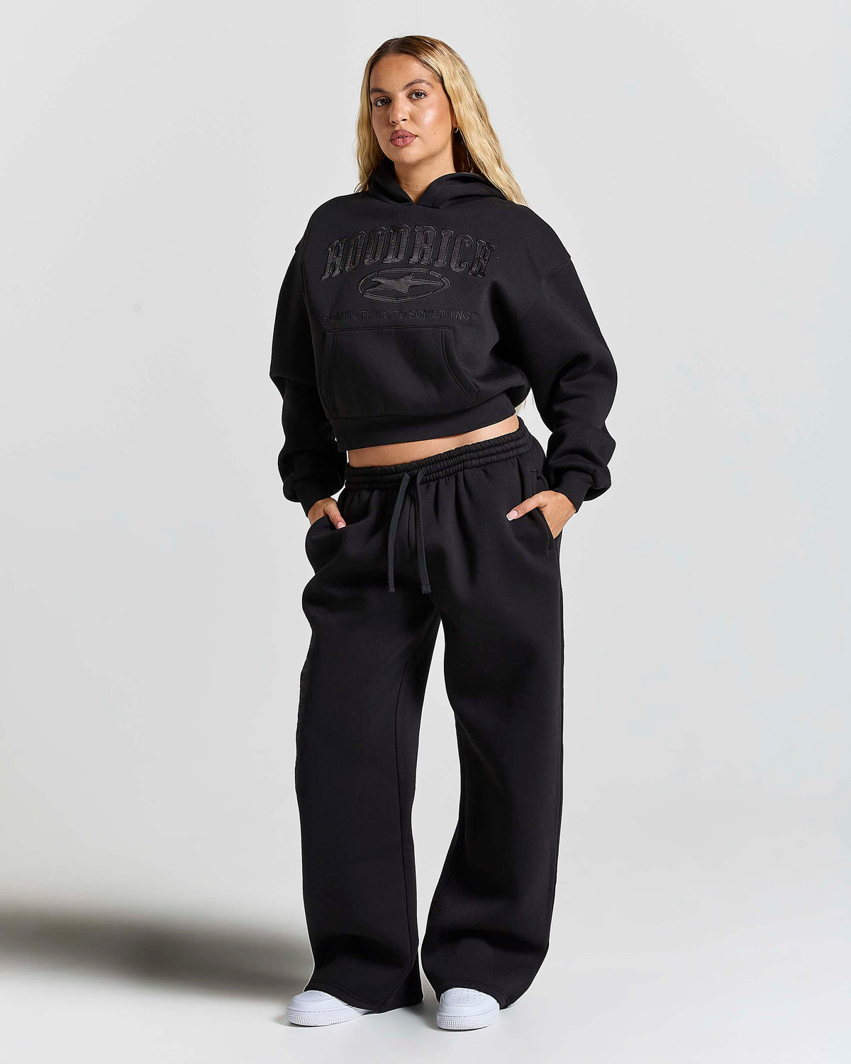 A woman modeling a black cropped hoodie and matching wide-leg sweatpants set. The hoodie features a tonal black graphic on the chest with the word 'HOODRICH' and a horse logo.