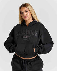 A woman modeling a black cropped hoodie with tonal black 'HOODRICH' text and logo across the chest, paired with matching black sweatpants, against a light gray studio background.