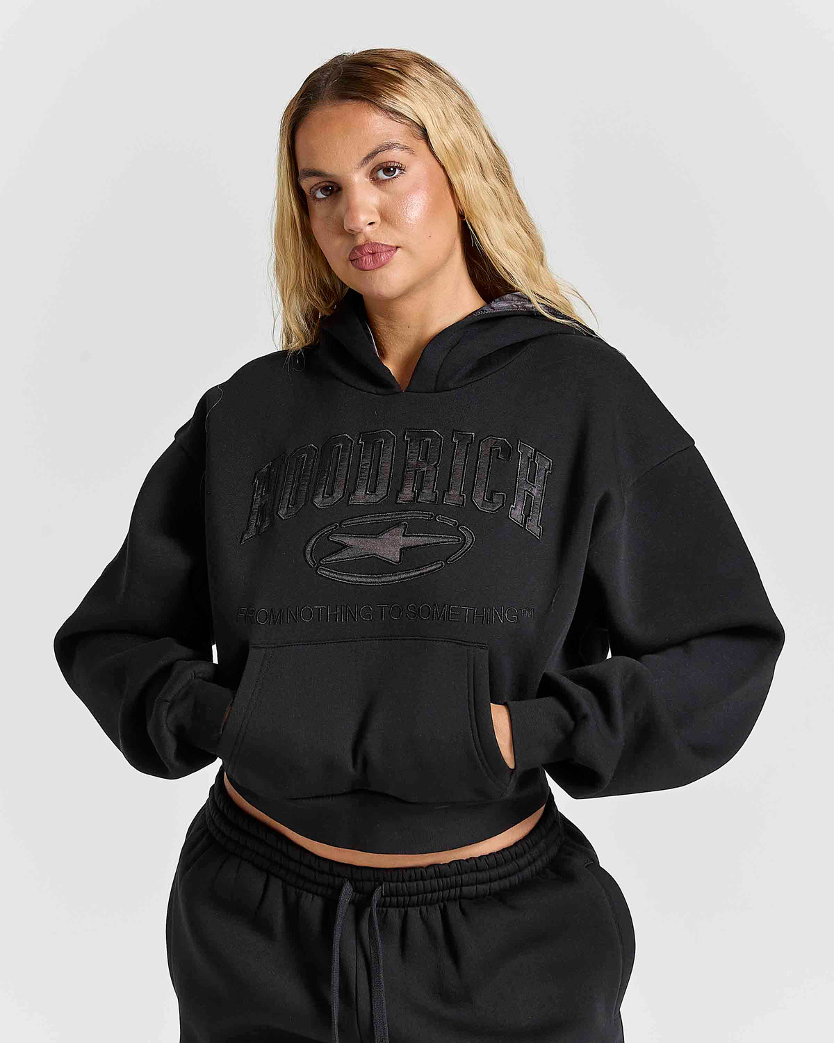 A woman modeling a black cropped hoodie with tonal black 'HOODRICH' text and logo across the chest, paired with matching black sweatpants, against a light gray studio background.