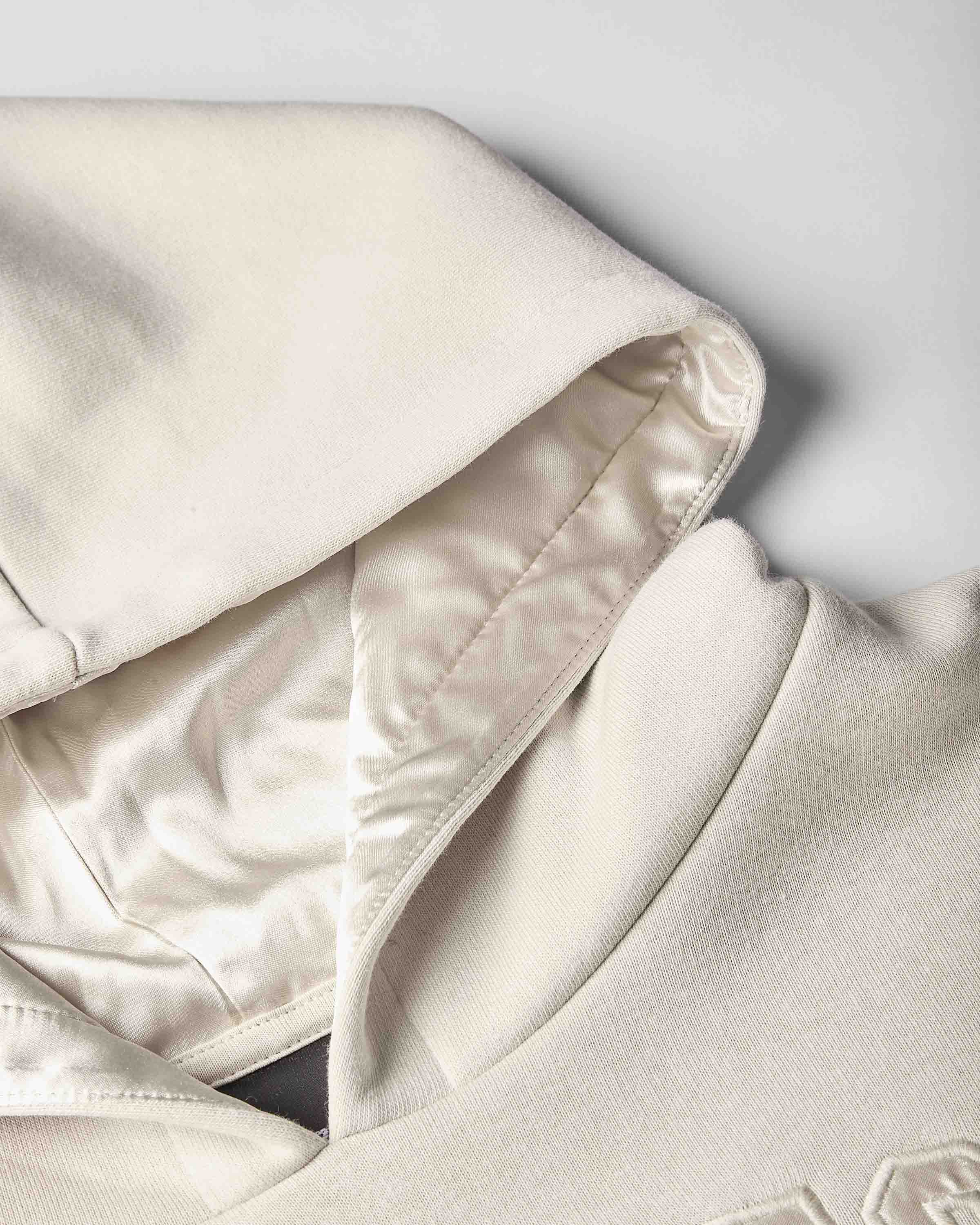 Close-up of a cream-colored hoodie showing the interior satin lining of the hood and collar, featuring a smooth satin-like inner fabric and a textured knit outer fabric.