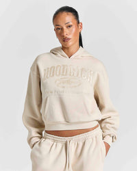 A model wearing a cream-colored cropped hoodie with 'HOODRICH' embossed on the chest, paired with matching jogger pants, against a light gray studio background.
