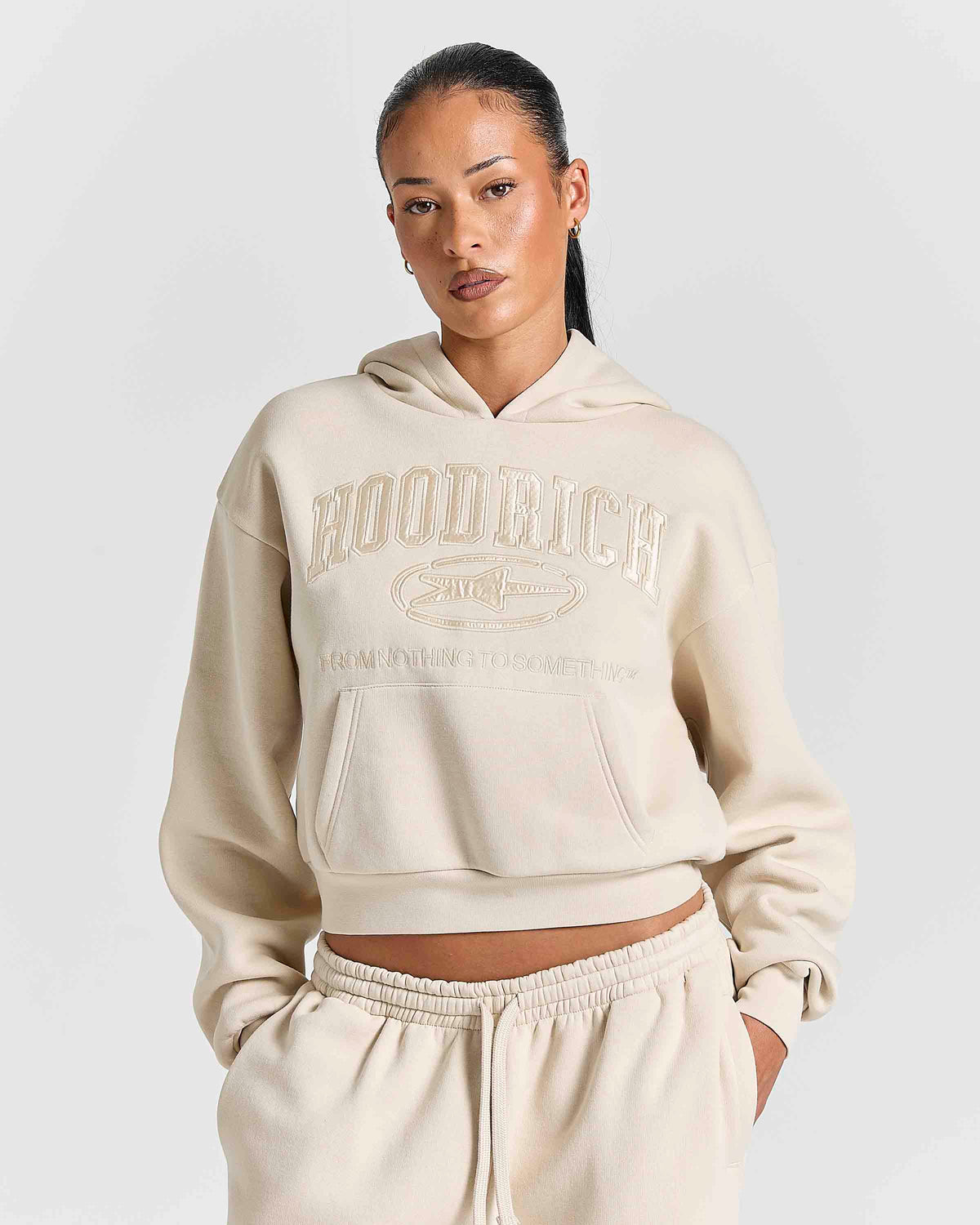 A model wearing a cream-colored cropped hoodie with 'HOODRICH' embossed on the chest, paired with matching jogger pants, against a light gray studio background.