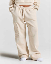A person wearing cream-colored wide-leg sweatpants with a vertical tonal 'HOODRICH' logo on the left leg, paired with white sneakers against a light gray studio background.
