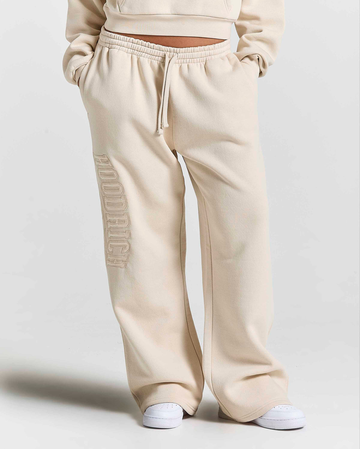 A person wearing cream-colored wide-leg sweatpants with a vertical tonal 'HOODRICH' logo on the left leg, paired with white sneakers against a light gray studio background.