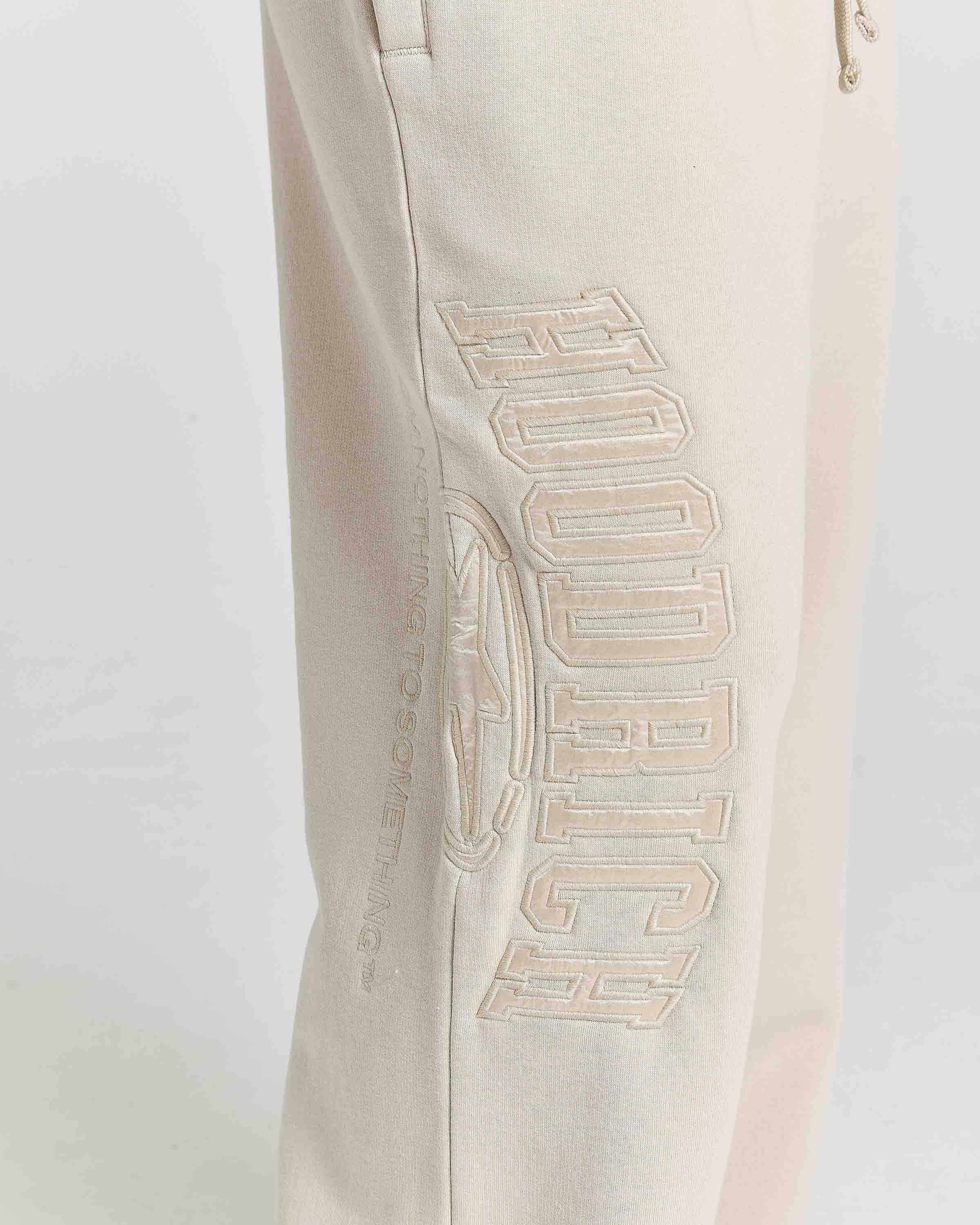Close-up of a light beige sweatpants leg featuring a large, tonal embroidered graphic of the word 'HOODRICH' with a circular logo emblem on the side.
