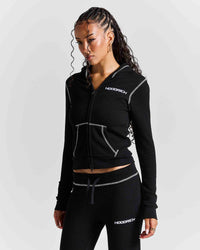 A woman modeling a black zip-up hoodie and matching black joggers with white contrast stitching and white HODGICH logos, photographed against a light gray studio background.