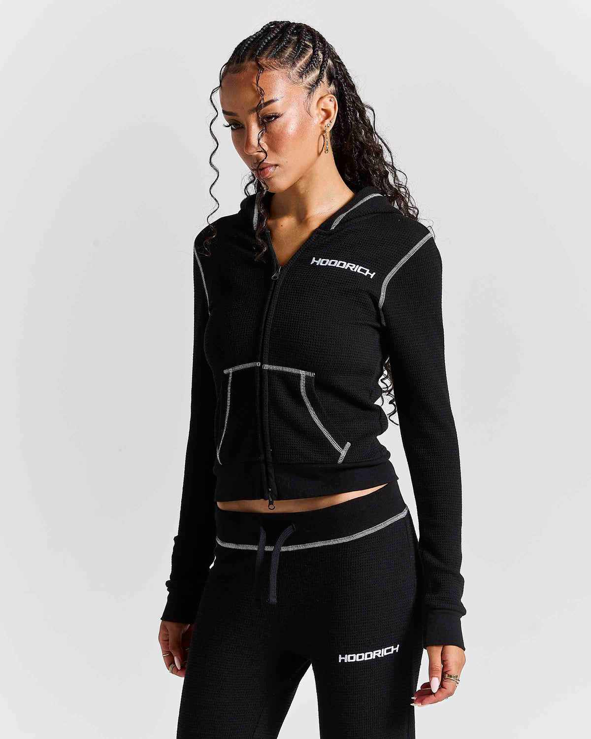 A woman modeling a black zip-up hoodie and matching black joggers with white contrast stitching and white HODGICH logos, photographed against a light gray studio background.